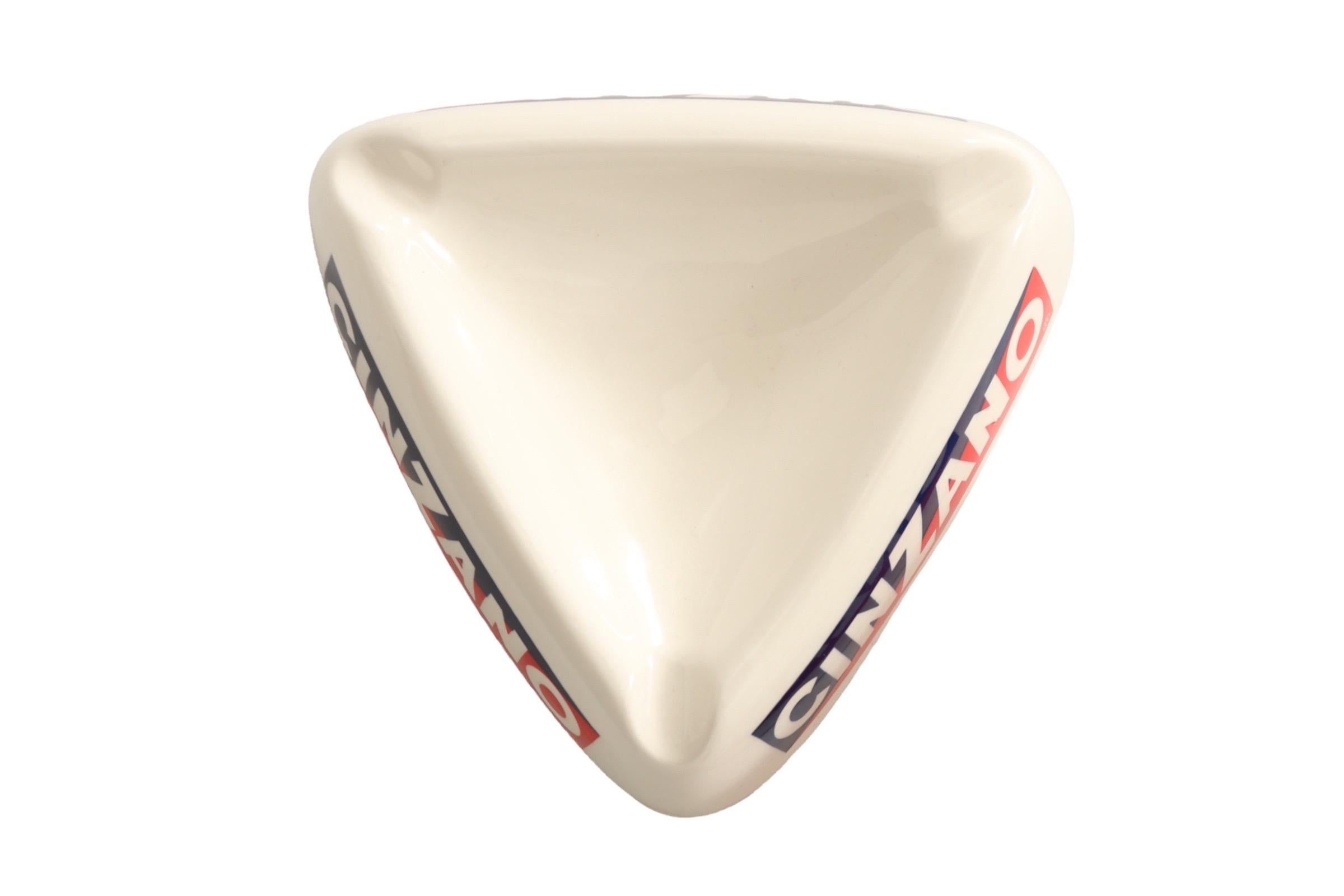 A triangular ceramic ashtray with the distinctive Cinzano logo in red, white and blue on all three sides, and a cigarette rest at each corner.
 