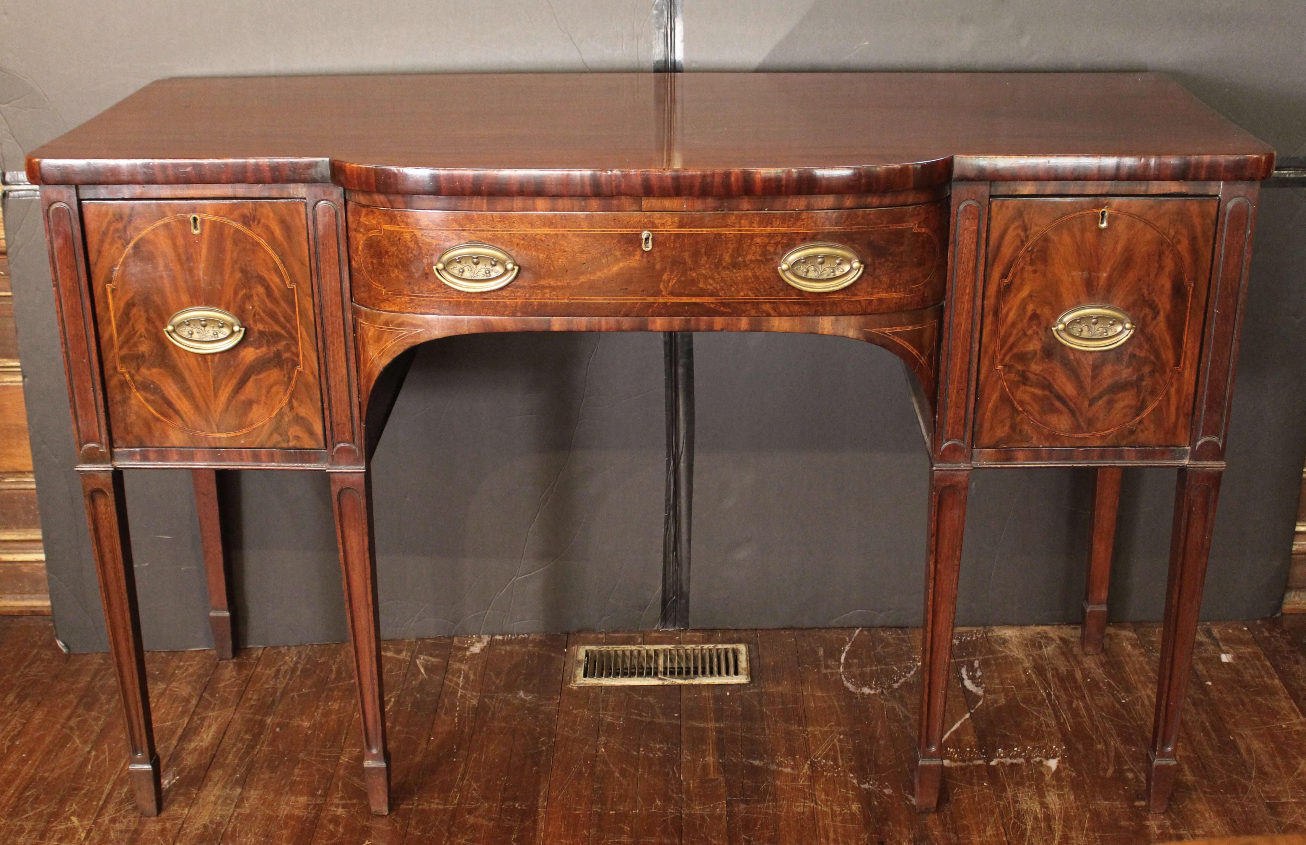 Circa 1780 George III sideboard, English. Mahogany with oak & pine secondary wood. Bow center breakfront form. Raised on straight, tapered legs with elongated oval fluting carrying on the carved motif flanking the drawers, ending in spade feet.