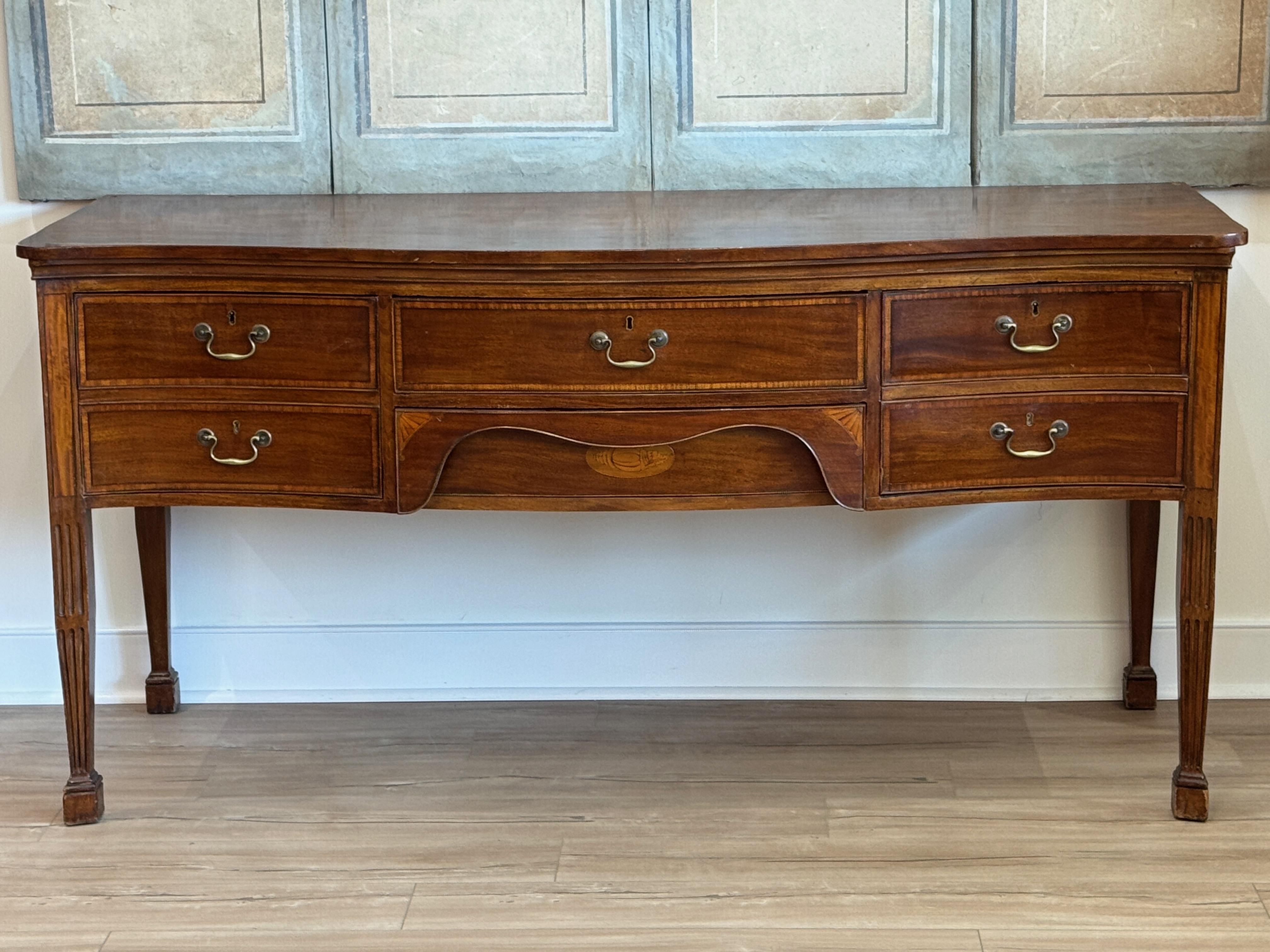 A George III period sideboard, circa 1795, in richly figured mahogany with satinwood banding and a serpentine front. The case features a central fitted drawer flanked by deep drawers and a shaped apron, all resting on fluted square tapering legs.