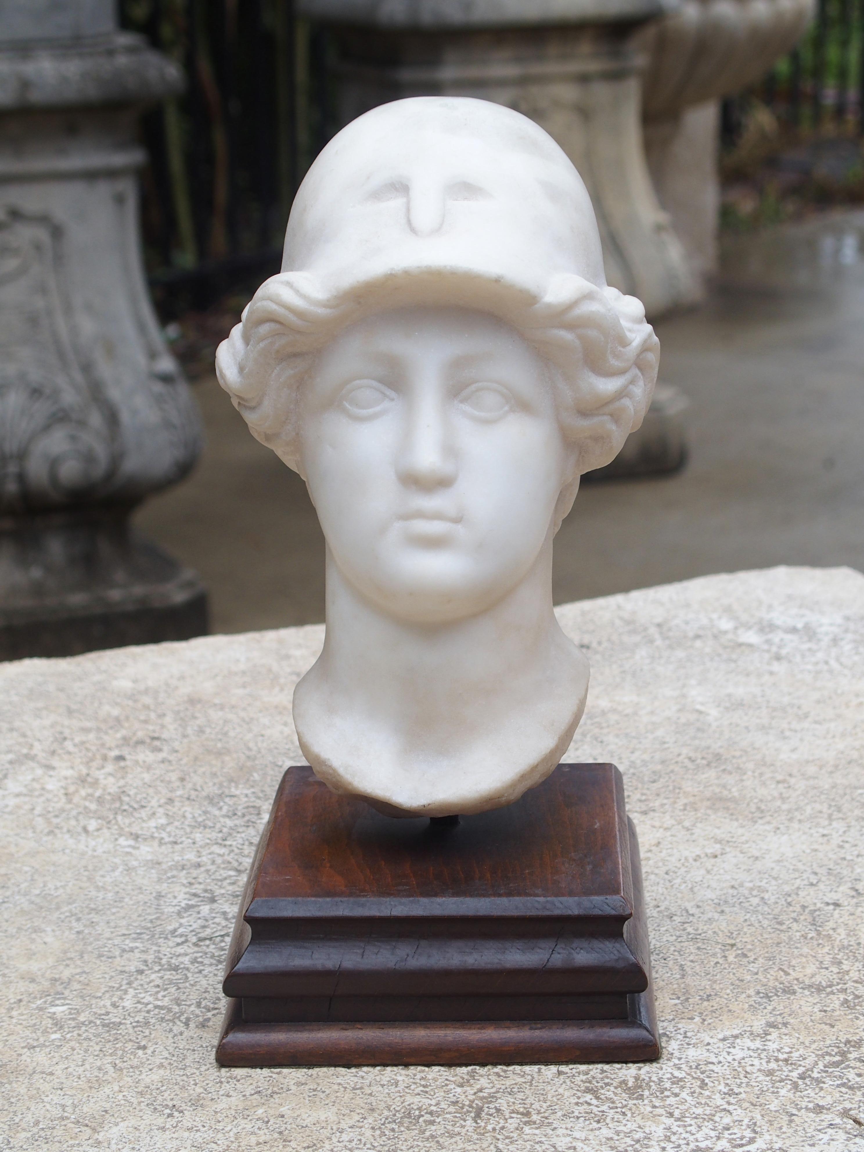 This is an antique Italian carved marble head of a young man in a Greco/Roman helmet on a wooden base. The sculptor has adeptly portrayed his countenance as wide eyed innocence. The marble head rests on the wooden base by means of a small iron rod