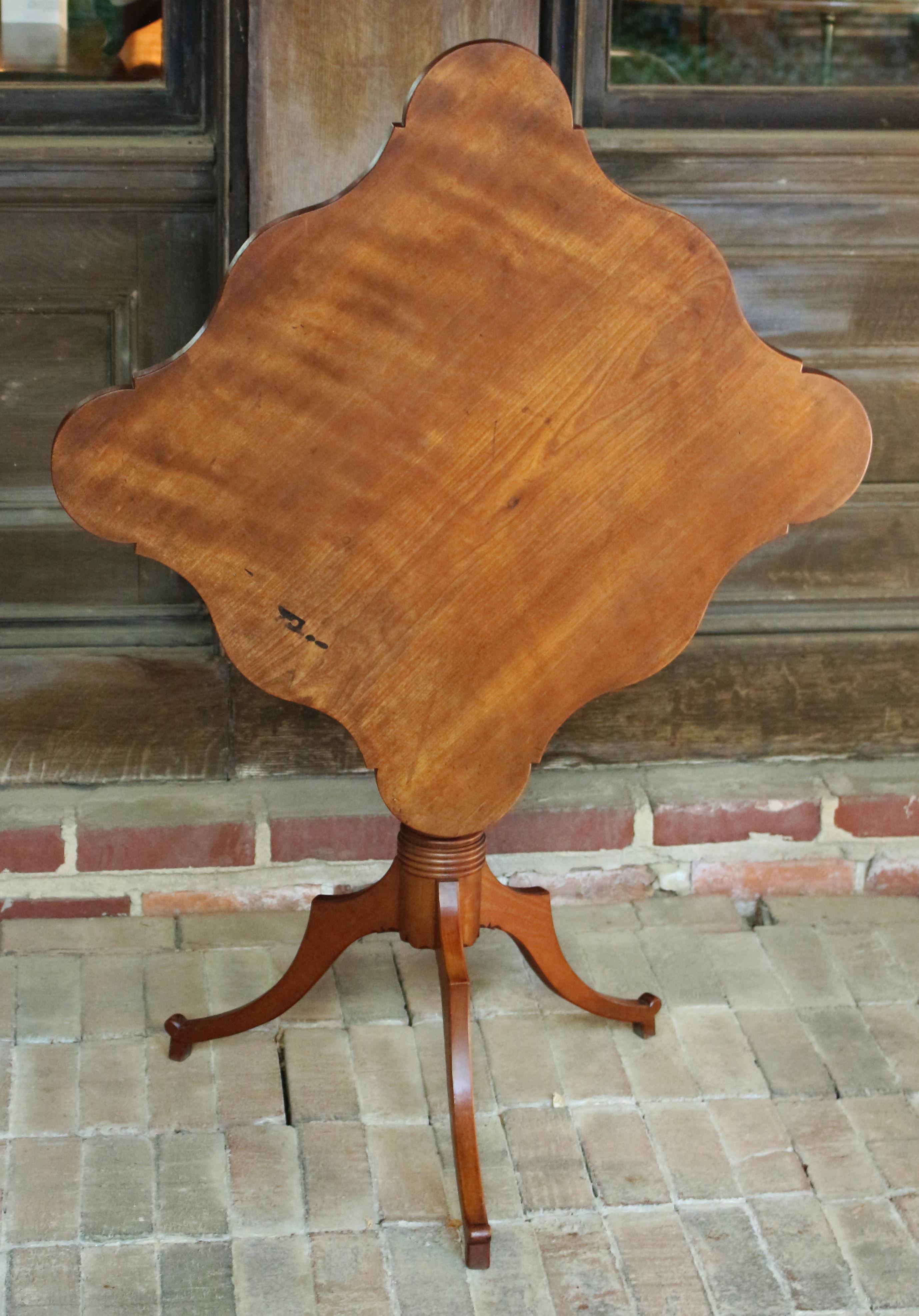 c.1820-30 American tilt-top tea table of cherry wood. Lovely shaped top, possibly done later or as an afterthought as it plays very well off the lovely shaped tripod pedestal base. Vasi-form and ring turned upright; shaped legs. Metal brace supports