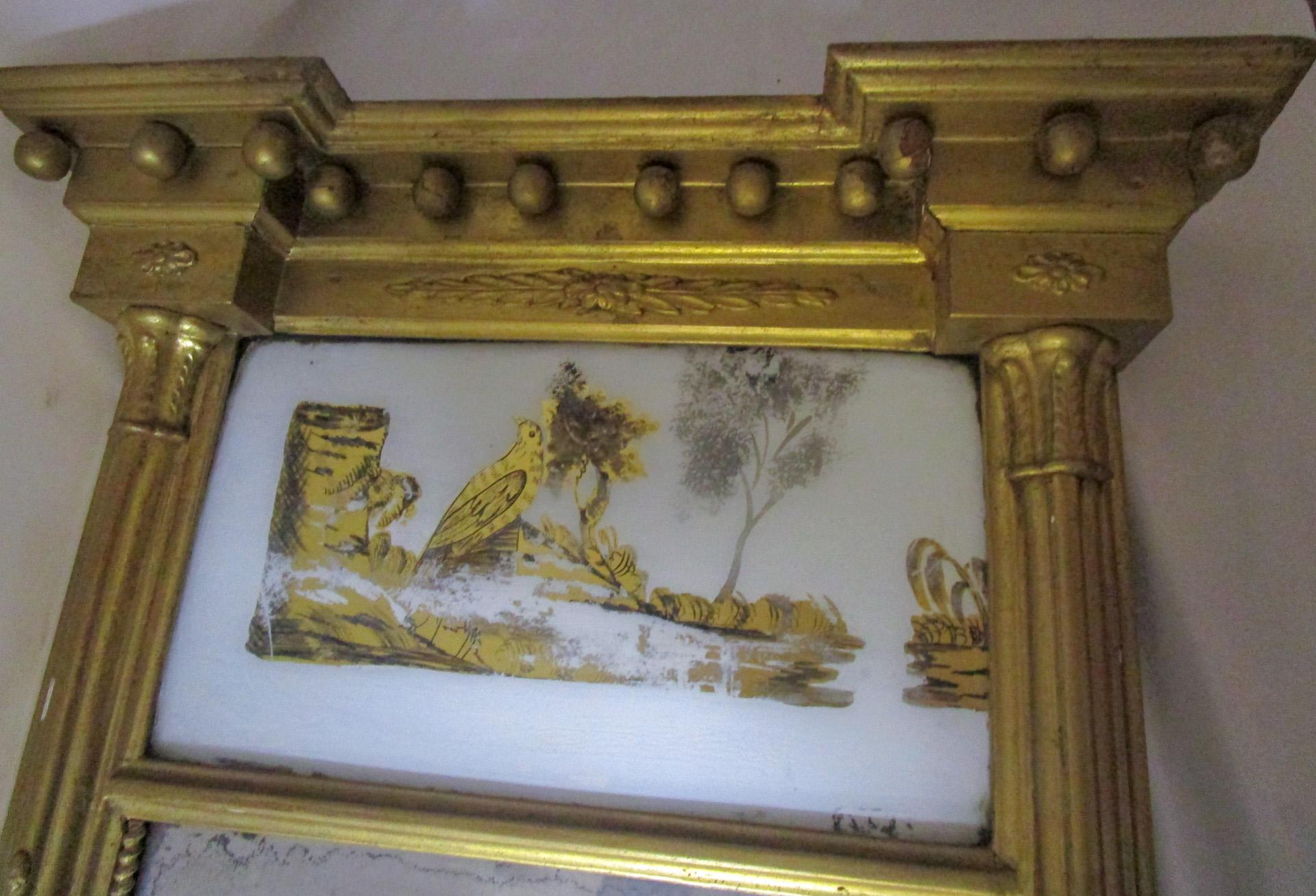 Circa 1820 giltwood architectural style looking glass with a reverse painted woodland scene eglomise tablet flanked with reeded columns, beading and appliques. Historian Rodric Blackburn writes that “the talent and some demand for such looking