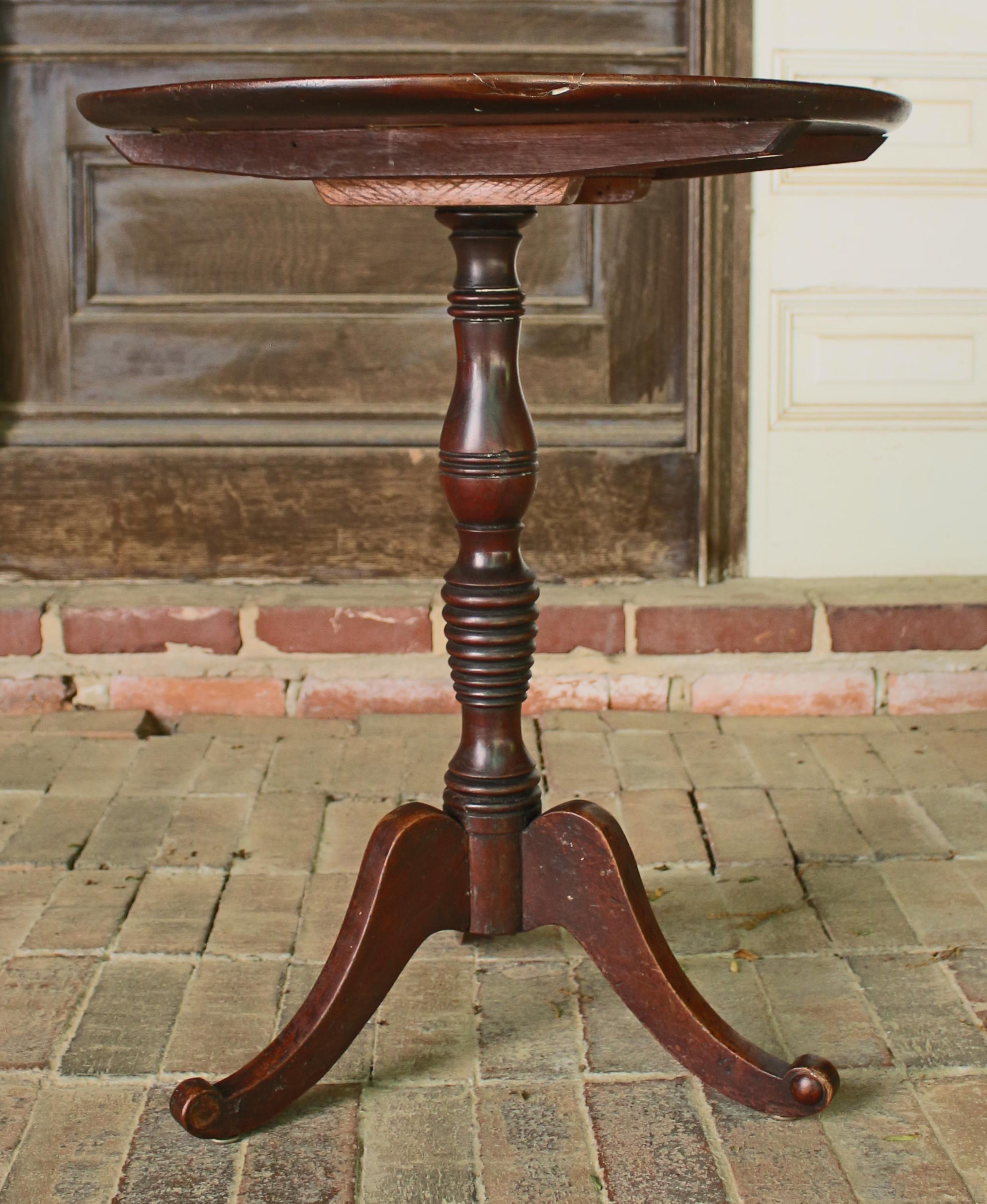 Late George III, c.1830, dish-top mahogany candlestand or small tea table with beehive turned standard raised on flat scrolled legs. Old top splits and repairs. Flipping mechanism partially restored. Split by one leg repairs. A charming, well used
