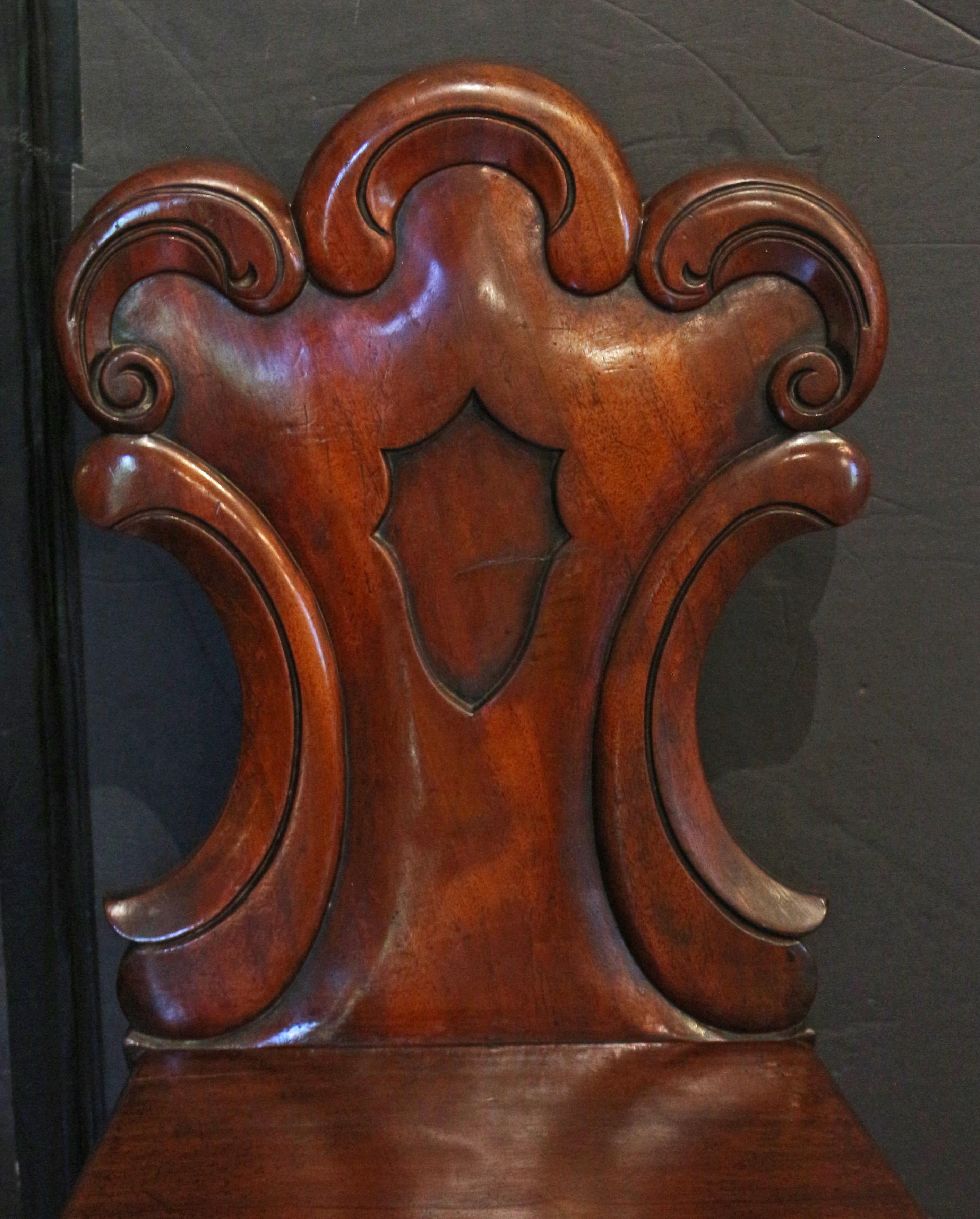 Circa 1860 pair carved hall chairs, English. Mahogany. Raised on boldly turned legs. The backs are of c-scrolled cartouche design centering a sculpted space for a crest. Great old finish, crusty in spots. Re-braced with blocks underneath the seat