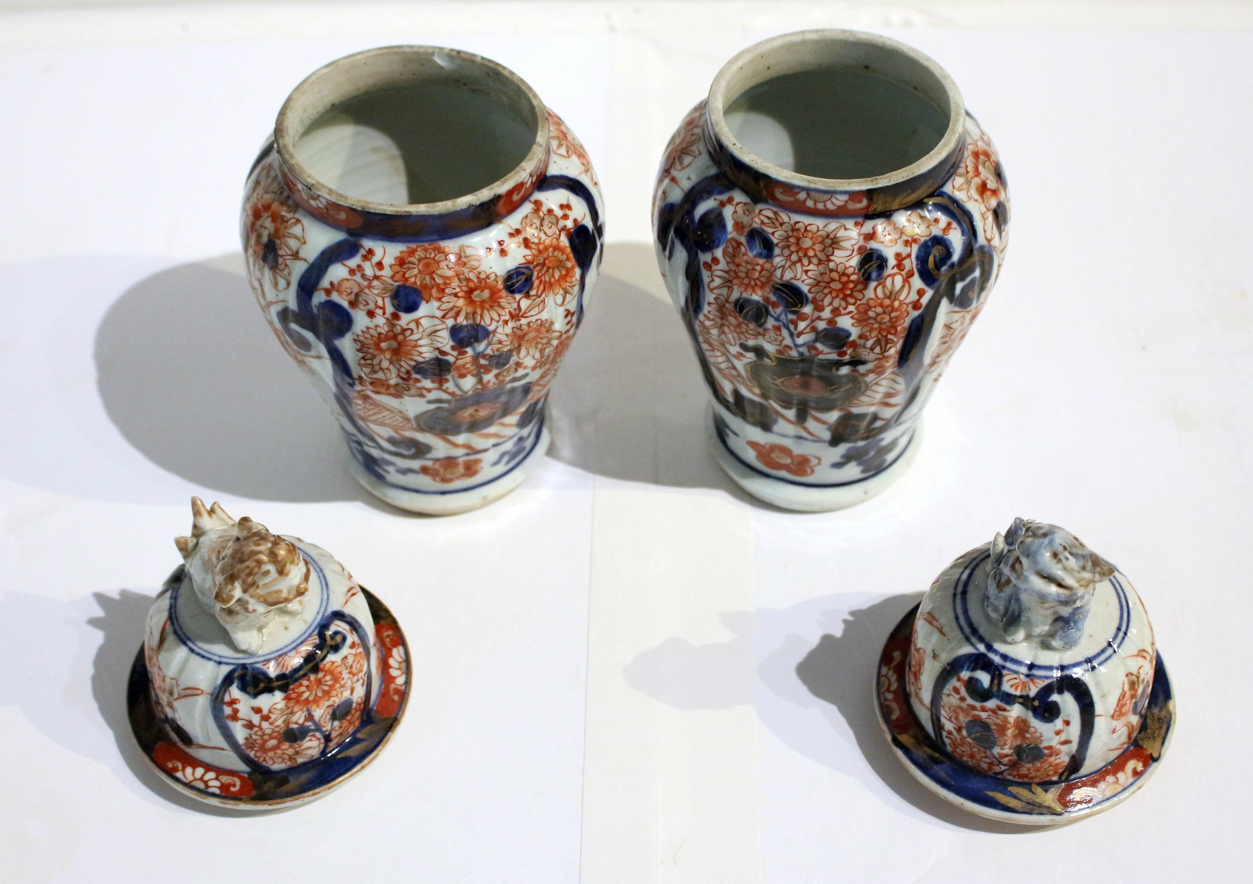 Circa 1860s pair of Imari covered vases, Japanese. Ribbed bodies. Well decorated with cartouches of exuberant floral vases. Foo dog finials - one with red-brown glaze highlights the other with blue. Interior rim chip to one. Both with tail tip