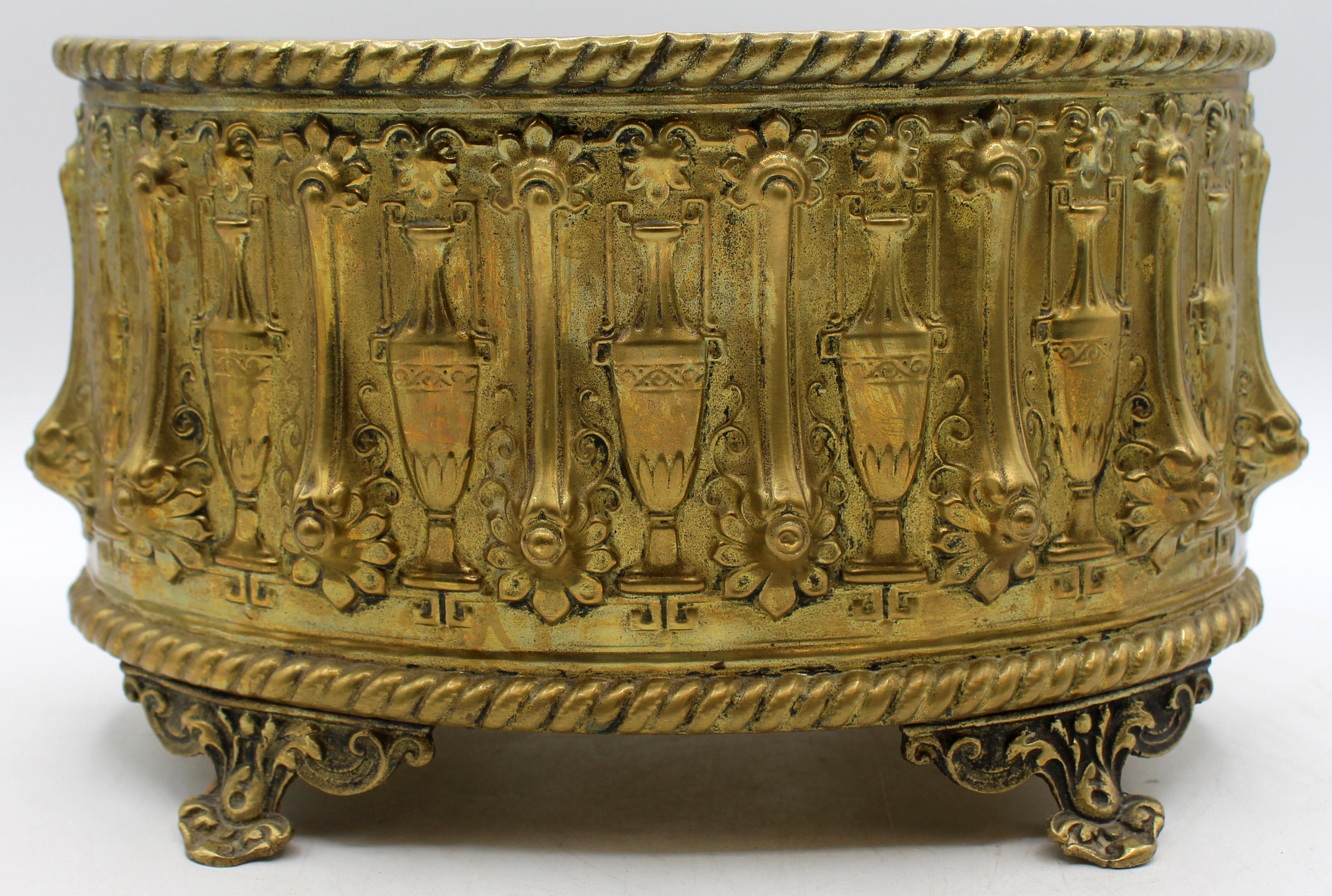 Circa 1870 oval repousse brass jardiniere, French or English. Original liner, as is. Gadrooned border & raised on 4 shaped floral feet. The body decorated with alternating classical urns & floret enhanced molded straps. Various minor dings & dents