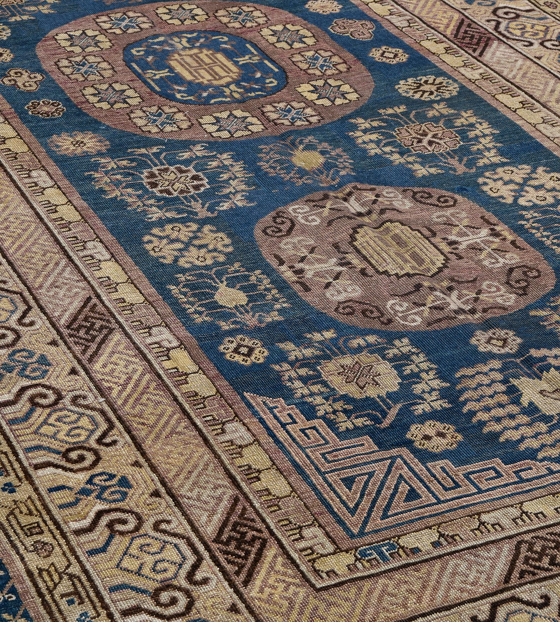 Dieser antike Khotan-Teppich aus der Zeit um 1880 hat ein tiefblaues Feld, das mit einer Vielzahl von Blütenköpfen und blättrigen Blumenmotiven um drei aubergine-lilafarbene Rondelle verstreut ist, das kleinere an jedem Ende mit einer zentralen,
