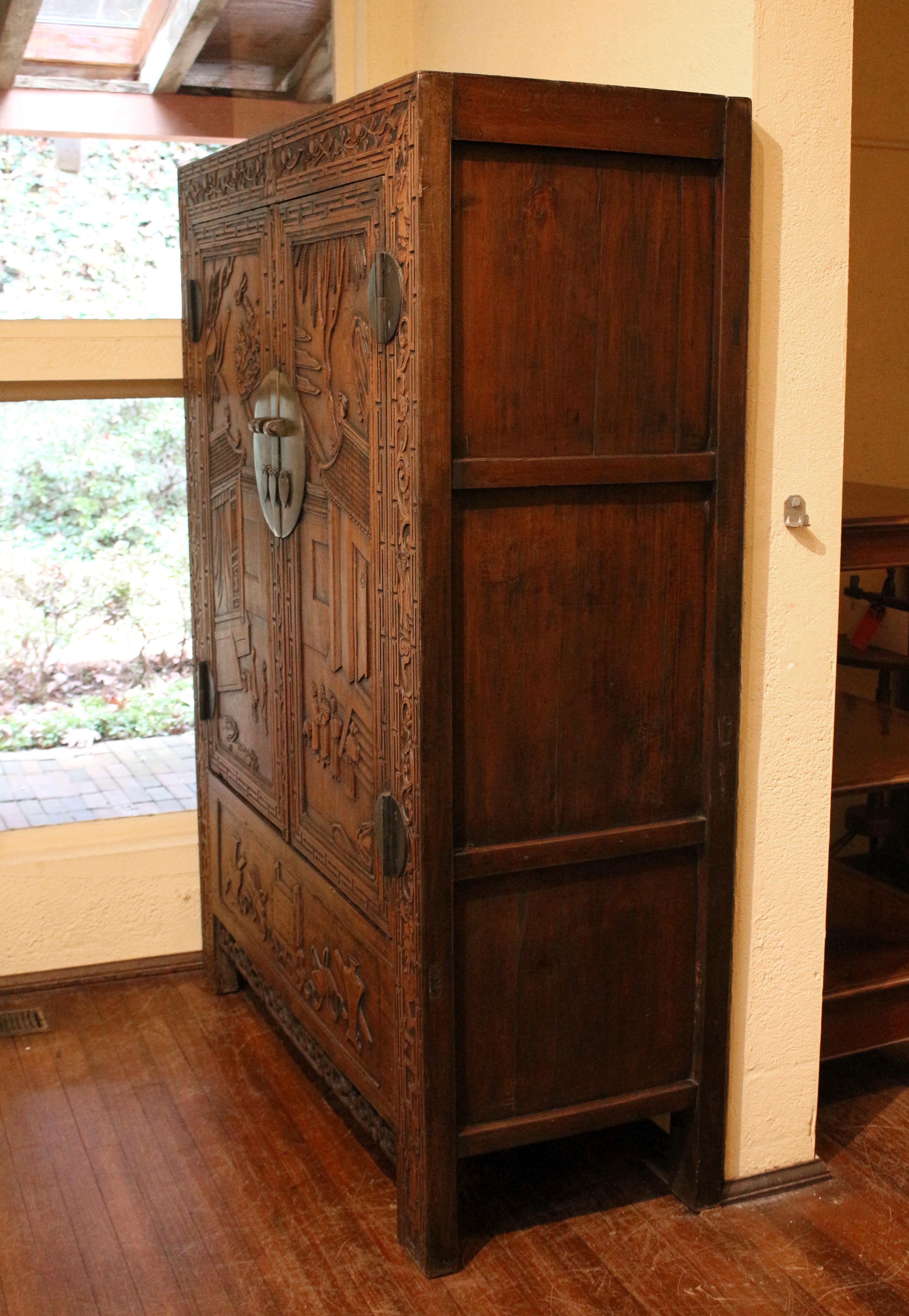 Circa 1880 Chinese carved wardrobe cabinet, Qing dynasty. Red stain on panelled sides. Extensively carved front. Perhaps for a marriage gift. Note the mythical figures on the roof and what seem to be gifts in the bottom frieze and in the arms of the