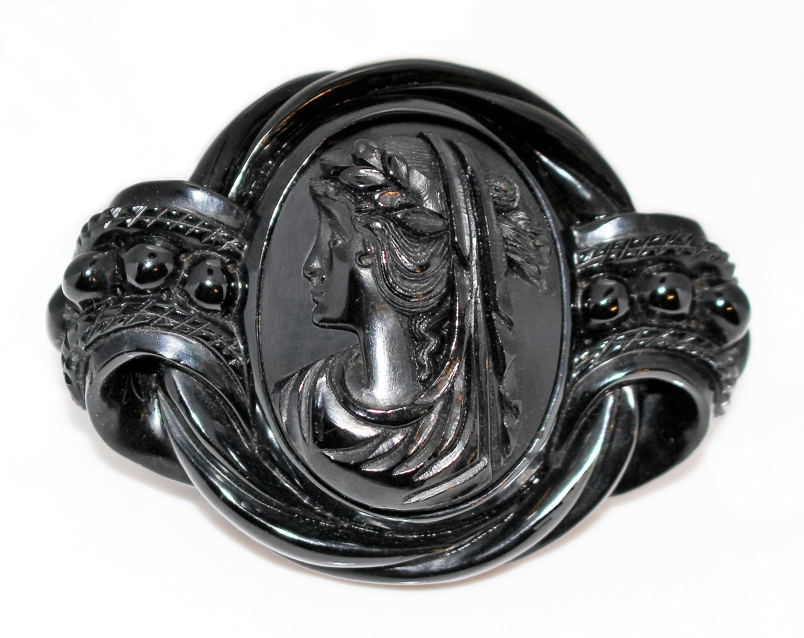 Circa 1880s Victorian Whitby jet brooch with an ornately carved raised swirl design surrounding the beautifully carved cameo center. Gold tone metal c-clasp closure and two small loops for attaching to chain or clothing.  It measures 2.7