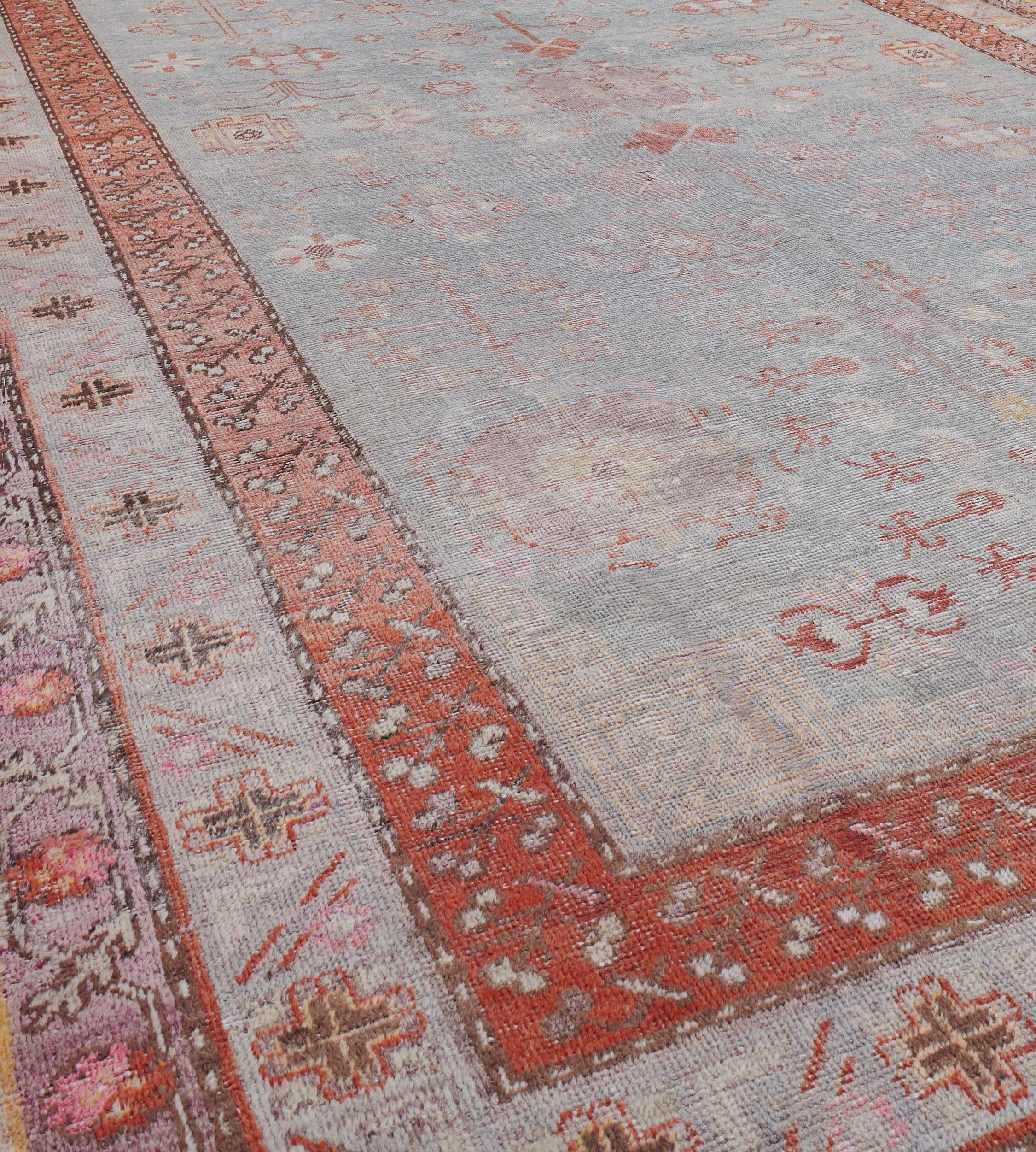 This antique, circa 1890, Khotan rug has a grey-blue field scattered with brick-red and light purple lozenges and angular hooked floral motifs, in a narrow sky-blue border of sandy-yellow and charcoal-black angular rosettes and cross-motifs