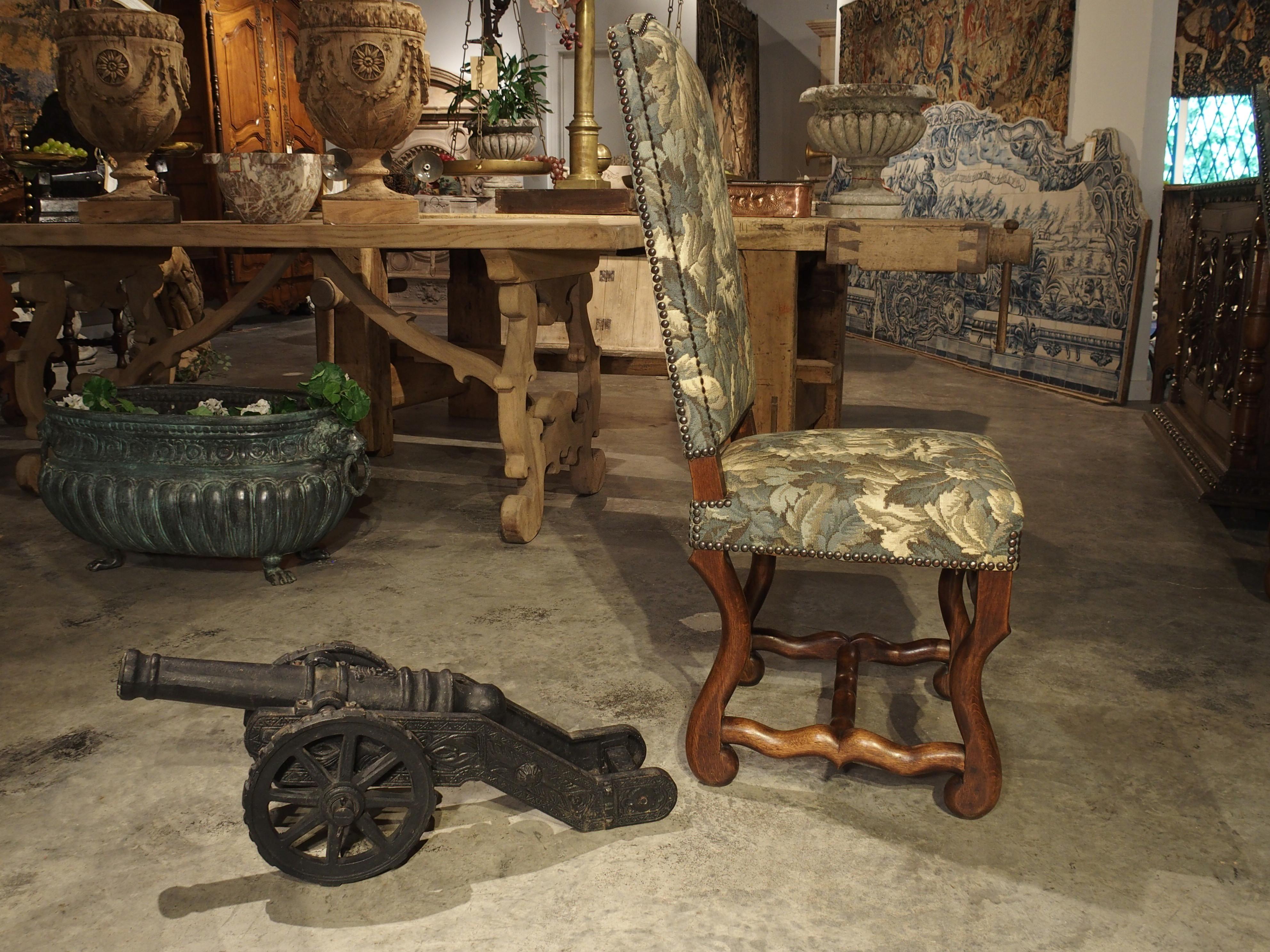 This 28 inch long black painted, cast iron cannon model has a coat of arms and foliate, floral and geometric patterns along its wheels and frame. The wheels have raised rectangular iron treads, and they are held to the axles by small metal pins.