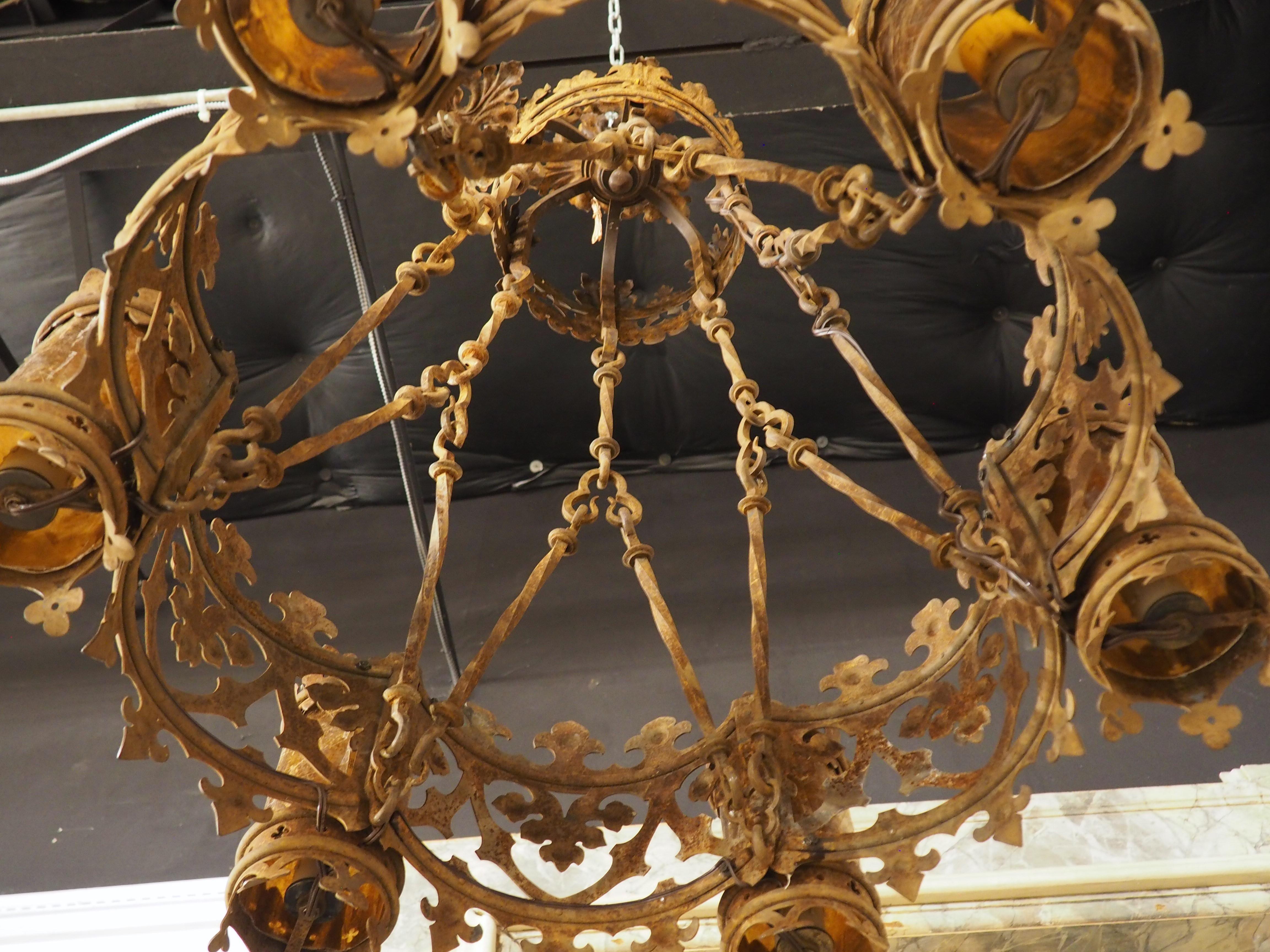 Bel exemple du style gothique, datant de la France d'environ 1900, ce lustre en fer forgé comporte six luminaires, chacun entouré d'un réflecteur ambré. Tous les luminaires s'inscrivent dans deux bandes de créneaux angulaires et d'arcades trilobées.