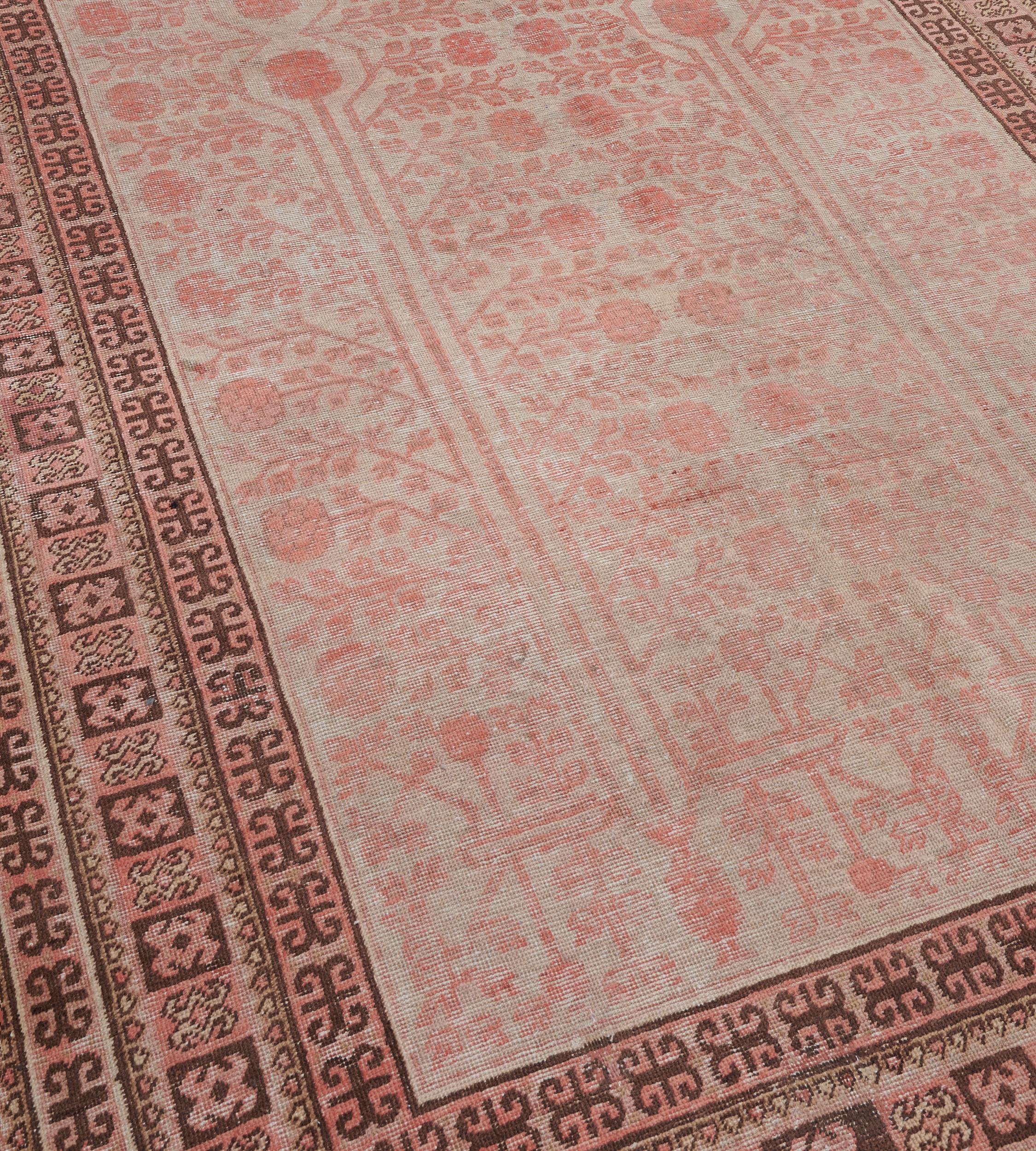 Ce tapis Khotan ancien, vers 1900, présente un champ gris avec un motif général de deux rangées de grenades rose-poussière et de vignes feuillues, dans une bordure jaune-sable de panneaux carrés roses-poussière et marron-chocolat reliés entre eux,