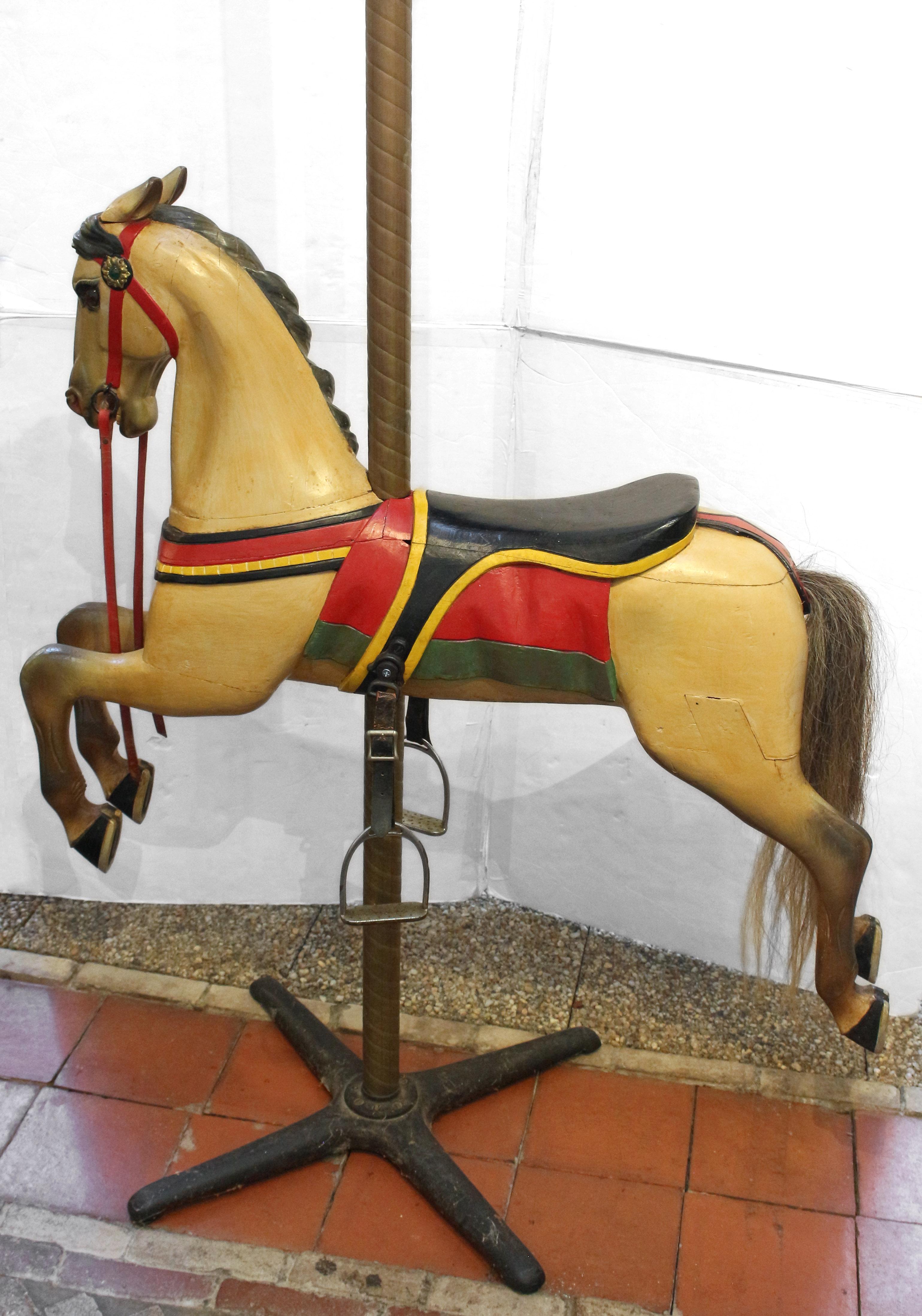 Circa 1900 carousel horse on original brass holder with iron stand, likely American. Carved wood - multiple pieces typical of antique figures. Apparently original paint. Steel stirrups. Red leather rein is broken. Leather stirrup straps are worn.