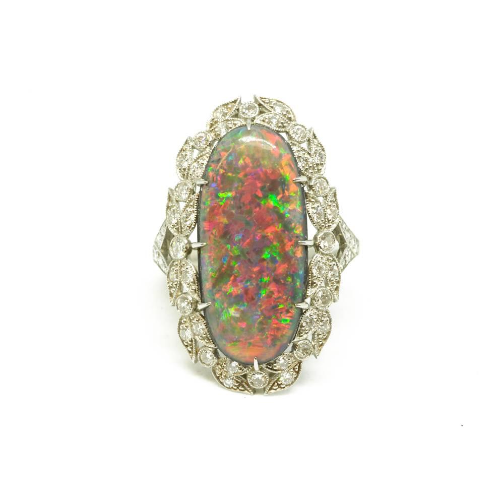 An oval black opal and diamond ring mounted in platinum. The elongated oval opal (7.87ct) has an intricate diamond set surround of foliate design with millegrain edging, and diamond set split shoulders. English circa 1910