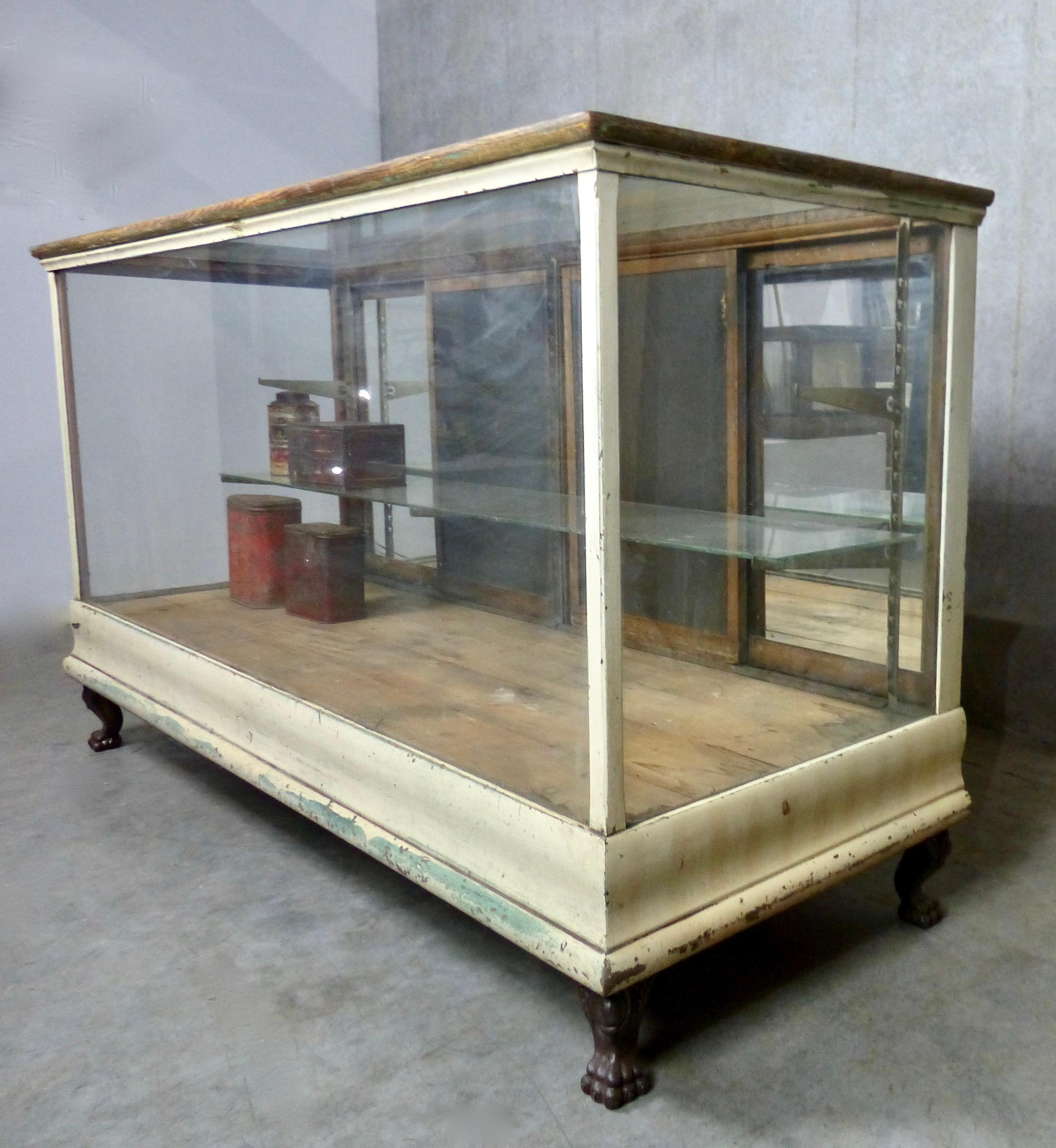 An antique mercantile cabinet elevated on cast copper feet. The cabinet features glass on three sides with some mirror on the rear sliding doors. The glass countertop (with beveled edges) has been beautifully aged over the past 100 years by eager