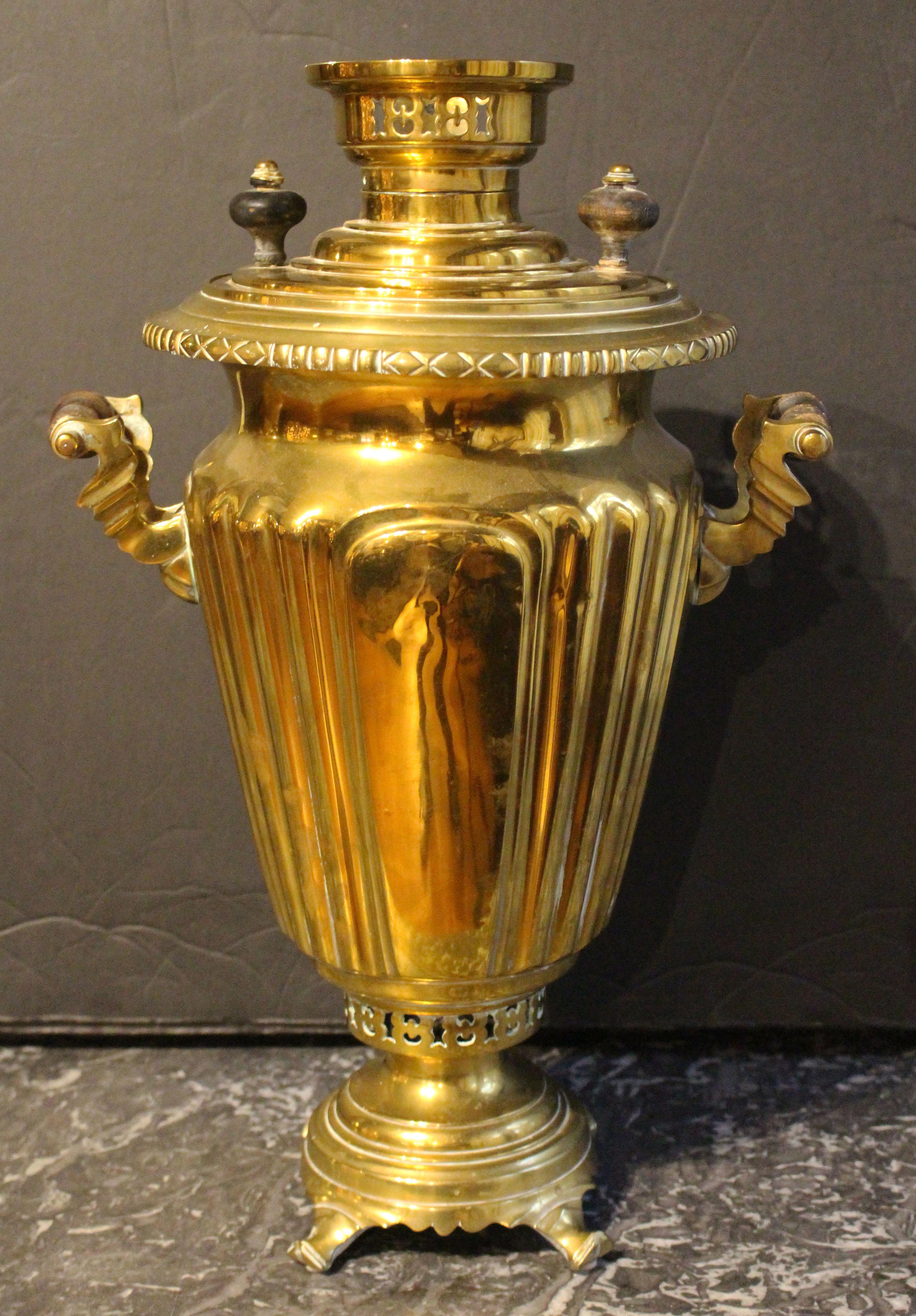 Samovar ruso de latón de hacia 1910. Elevado sobre patas con volutas; herrajes de latón fundido y madera torneada. Cuerpo estriado y cónico. Sellos de premio de las exposiciones de 1893 y 1900. Carece de tapones y plancha calefactora. Una