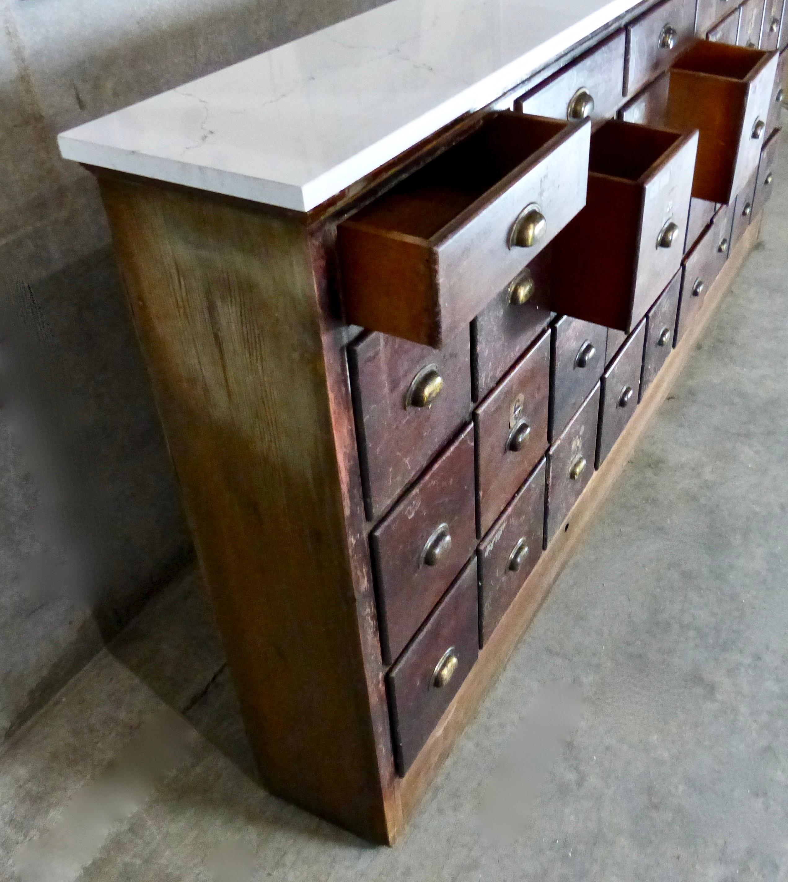 A multi-drawer, solid wood apothecary/hardware cabinet from circa 1910. Features include six flat drawers and 27 square drawers, marble top, and original brass hardware. The cabinet’s narrow depth makes it suitable for tight spaces like hallways or