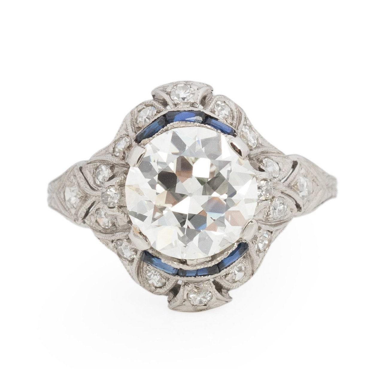 Behold this platinum Edwardian engagement ring of unparalleled beauty. Its tapered shanks bear intricate engravings resembling delicate leaves that gracefully unfurl as they ascend. The center gem is embraced by an openwork geometric halo adorned