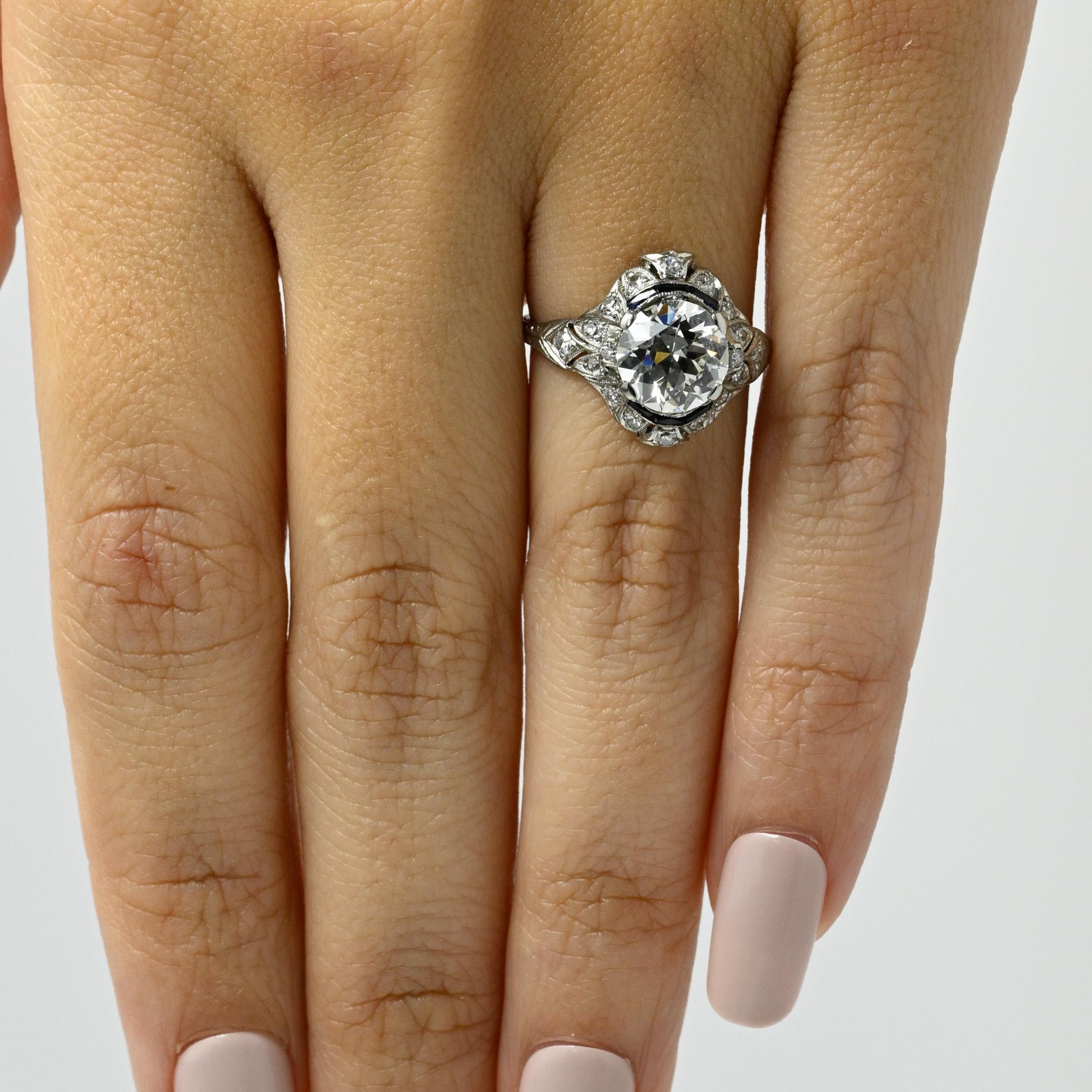 Behold this platinum Edwardian engagement ring of unparalleled beauty. Its tapered shanks bear intricate engravings resembling delicate leaves that gracefully unfurl as they ascend. The center gem is embraced by an openwork geometric halo adorned