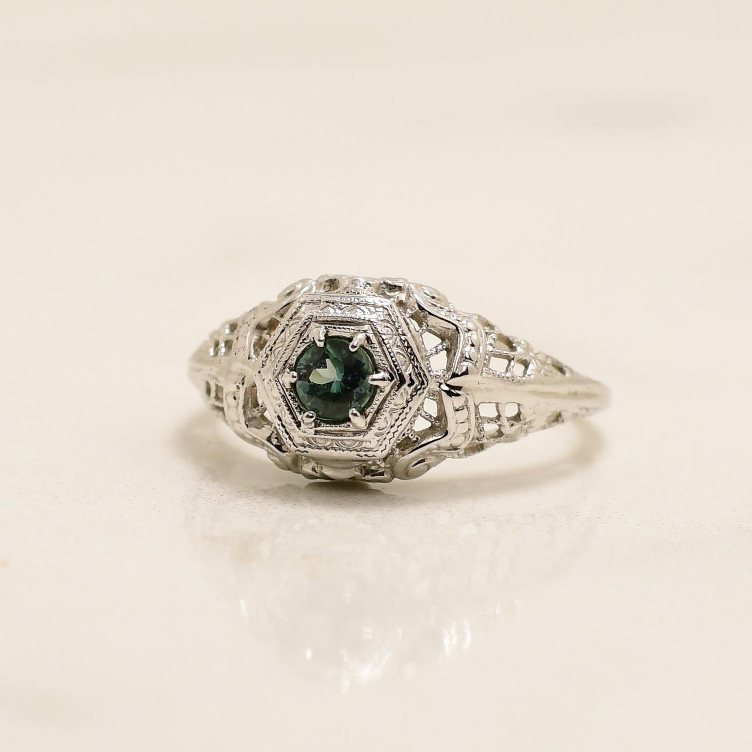 Behold the exquisite beauty of this Art Deco ring, a true masterpiece of design. Crafted with precision in 18K white gold, its filigree openwork evokes a charming floral essence. The lattice structure forms both the gallery and cathedral shanks,