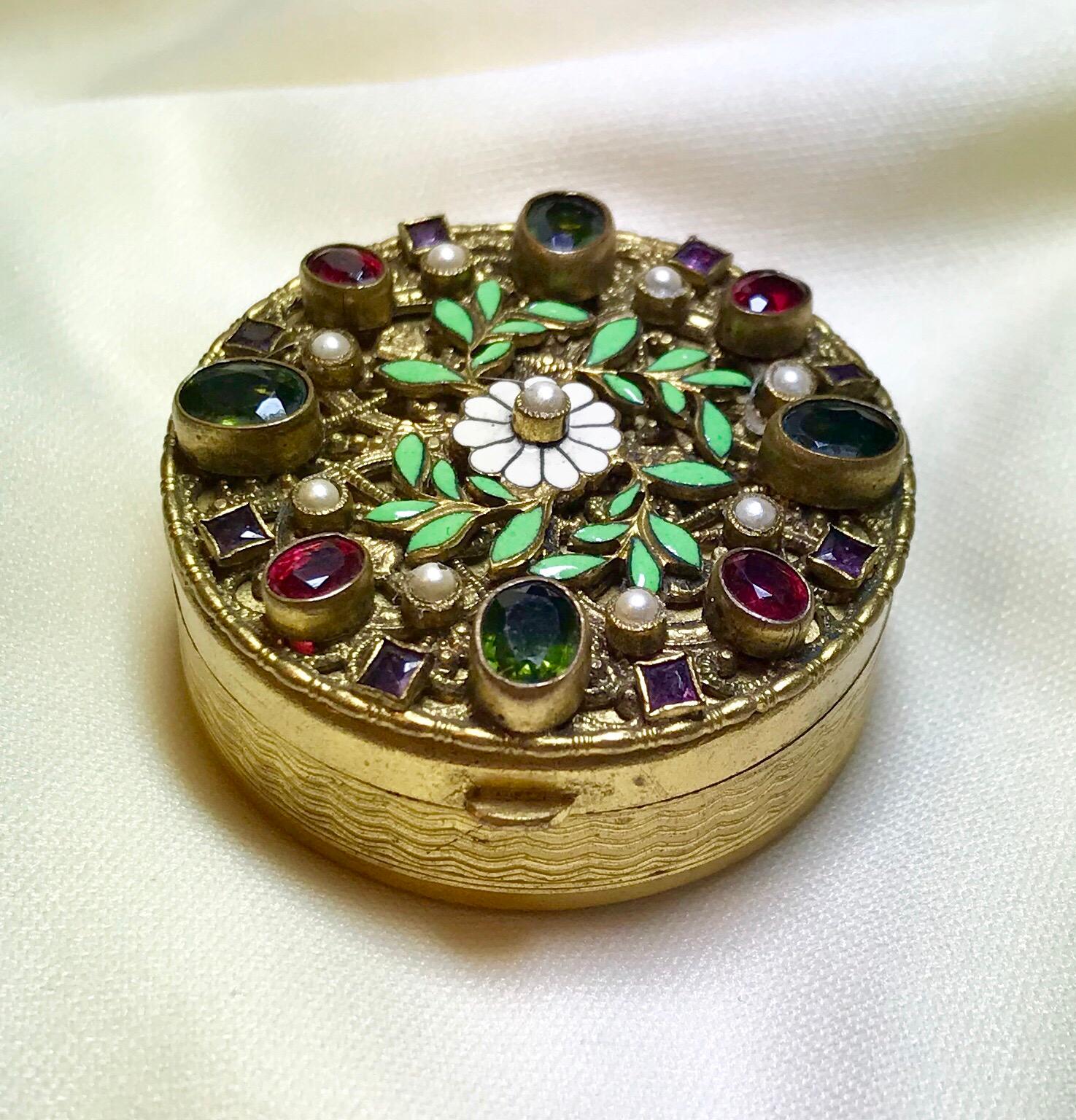 Circa 1920s gold tone Austrian powder compact with an engine turned design around the sides and on the bottom.  The top of the compact has an enameled floral and leaf design and is bezel set with faceted glass round and oval stones in green, red and