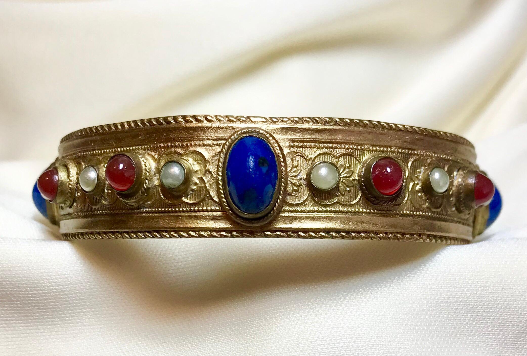 Circa 1920s plated brass bangle with a raised floral design and a rope motif edging.  It is bezel set with glass cabochons in shades of carnelian and lapis-blue and embellished with faux-pearls.  The interior measures 8.4