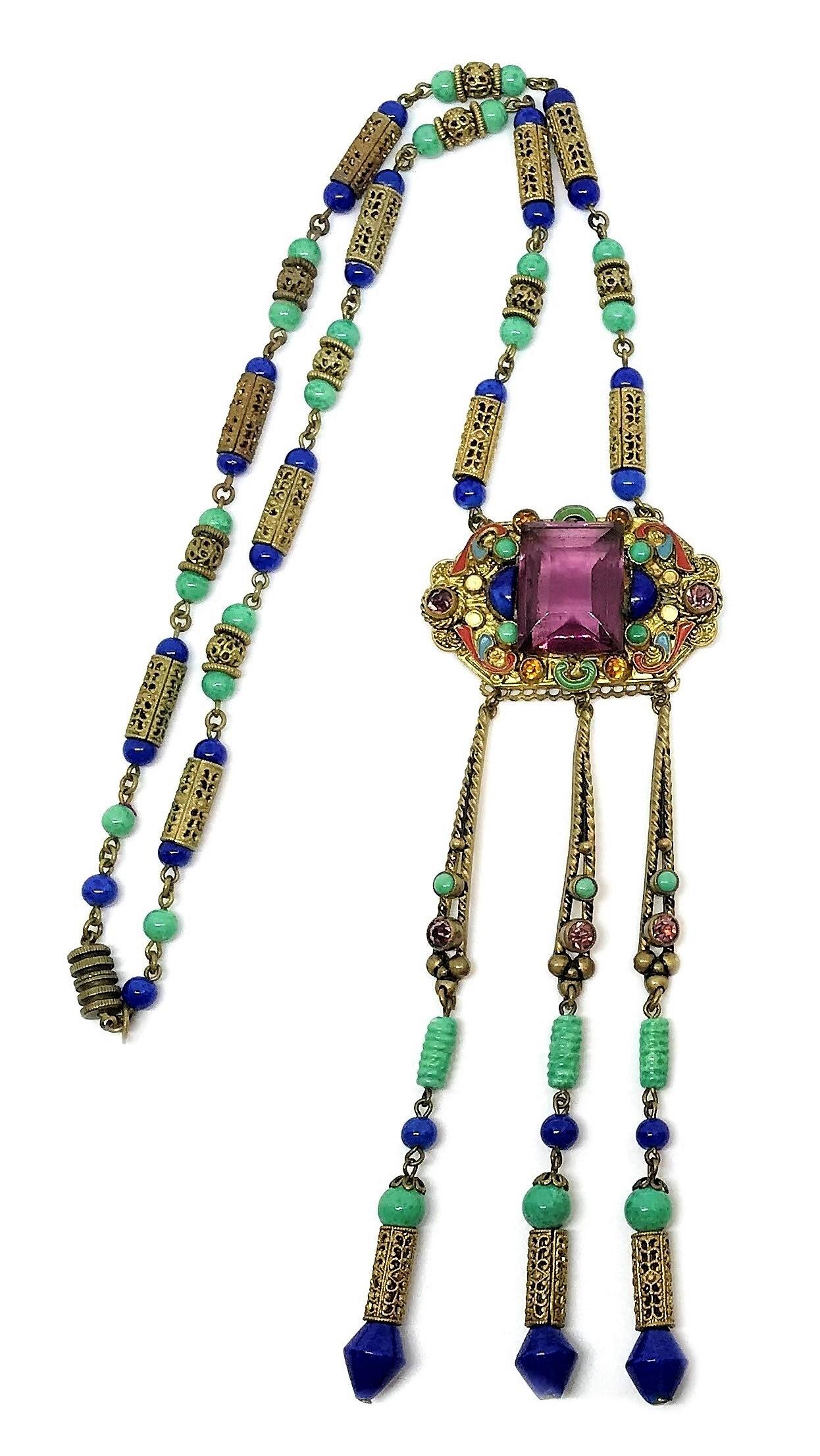 Circa 1920s to 1930s Czechoslovakian Egyptian Revival necklace with a brass pendant set with a large emerald cut, purple faceted glass stone. The pendent is embellished with multiple shape and color glass cabochons, red enameling and long jeweled