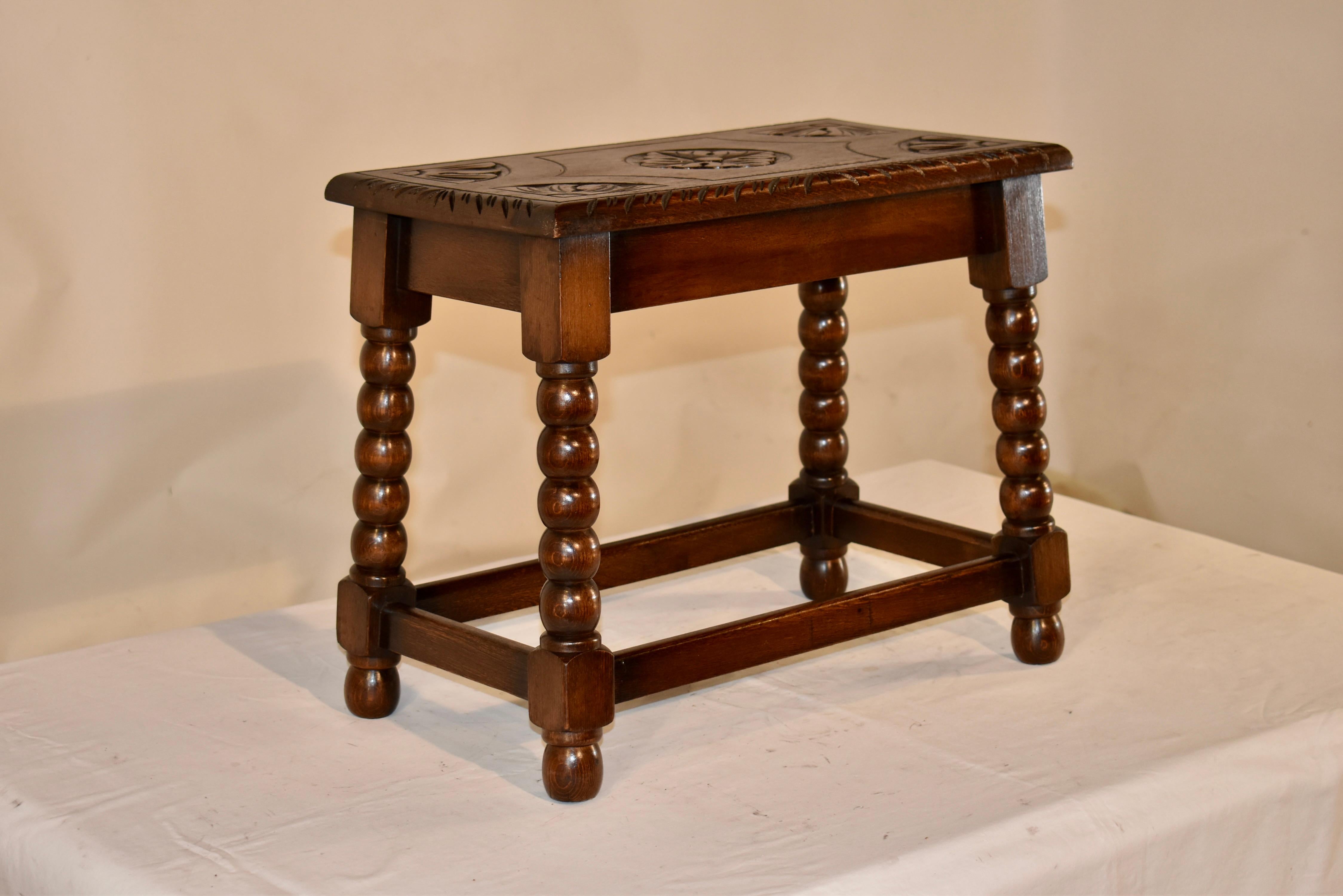 Circa 1920's English oak stool with a lovely hand carved design on the top, surrounded by a beveled and carved edge.  The apron is simple and the stool is supported on hand turned and splayed bobbin legs, joined by simple stretchers, and raised on
