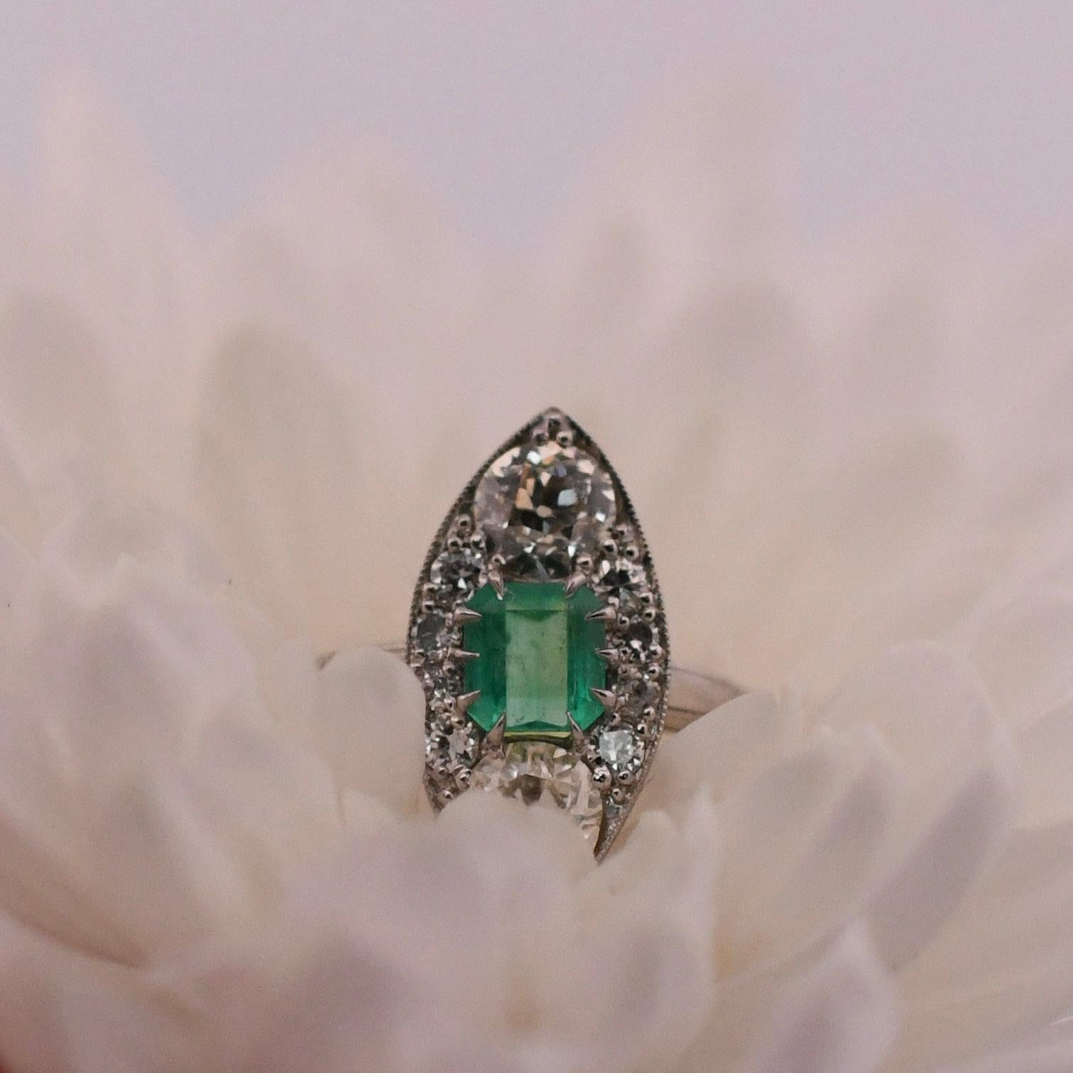 Versetzen Sie sich mit diesem exquisiten 3-Stein-Navette-Ring in die glamouröse Art-Déco-Ära. Zwei funkelnde Diamanten flankieren einen fesselnden kolumbianischen Smaragd in der Mitte. Das Design des Rings verkörpert die klaren Linien, die
