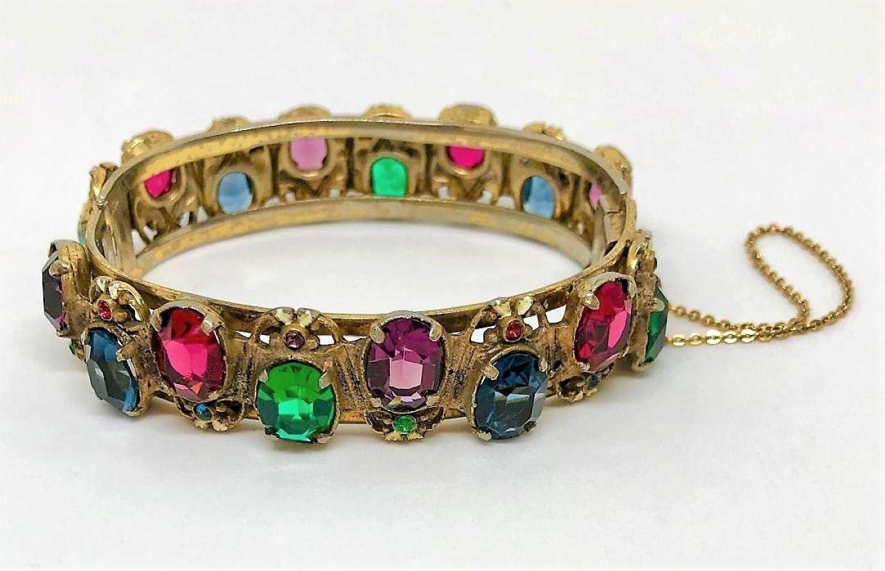 Circa 1930s gold tone plated metal hinged bangle prong set with large faceted oval glass stones and embellished with small stones and off-white enameling.  The open-back stones are ruby red, sapphire blue, emerald green and amethyst purple.  The