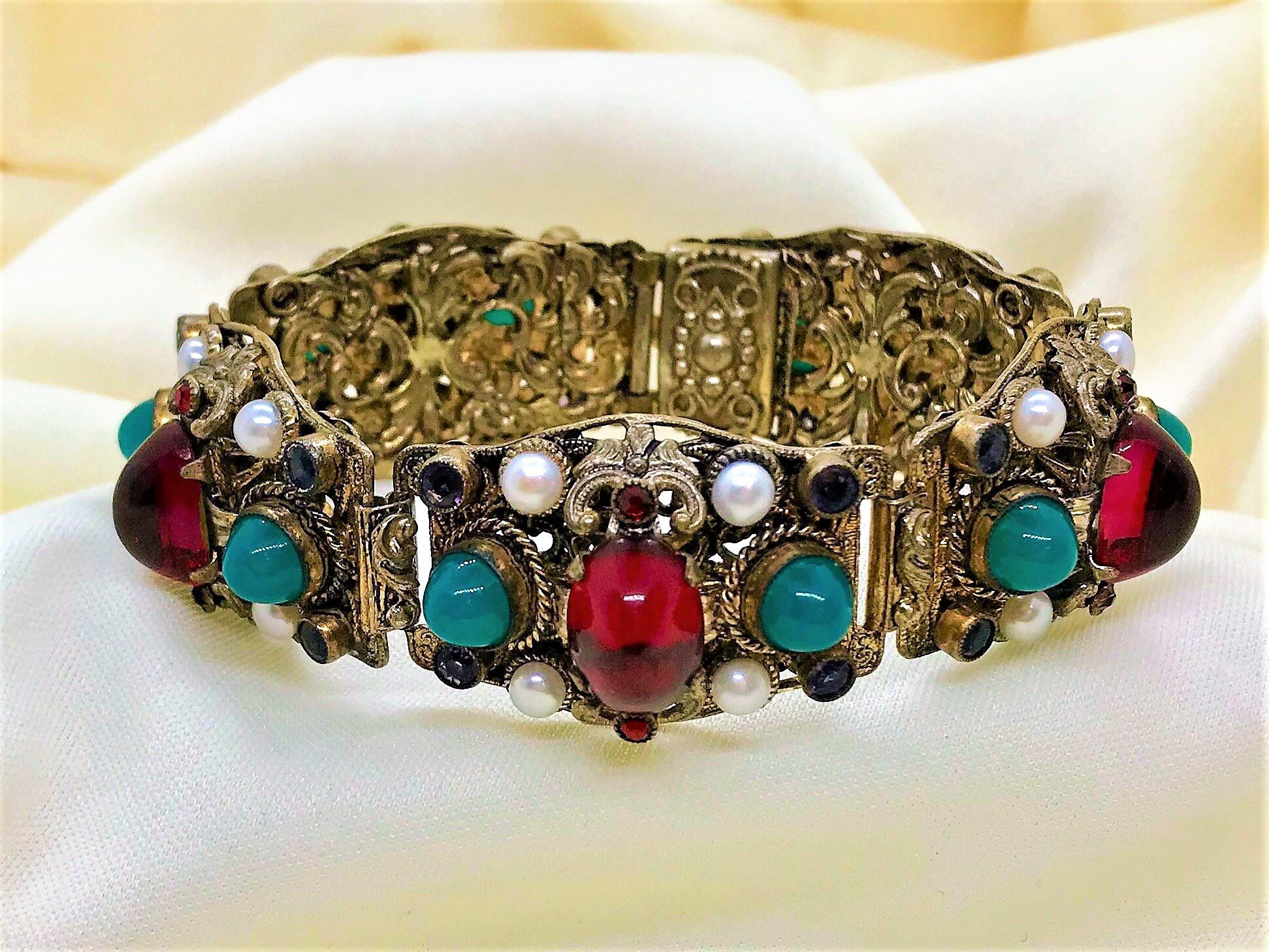 Circa 1940s to 1950s ornately designed, Austro Hungarian style, gold tone metal bracelet.  It is bezel and prong set with cultured pearls, ruby red and jade green glass cabochons as well as purple faceted glass stones.  The bracelet is 7.12