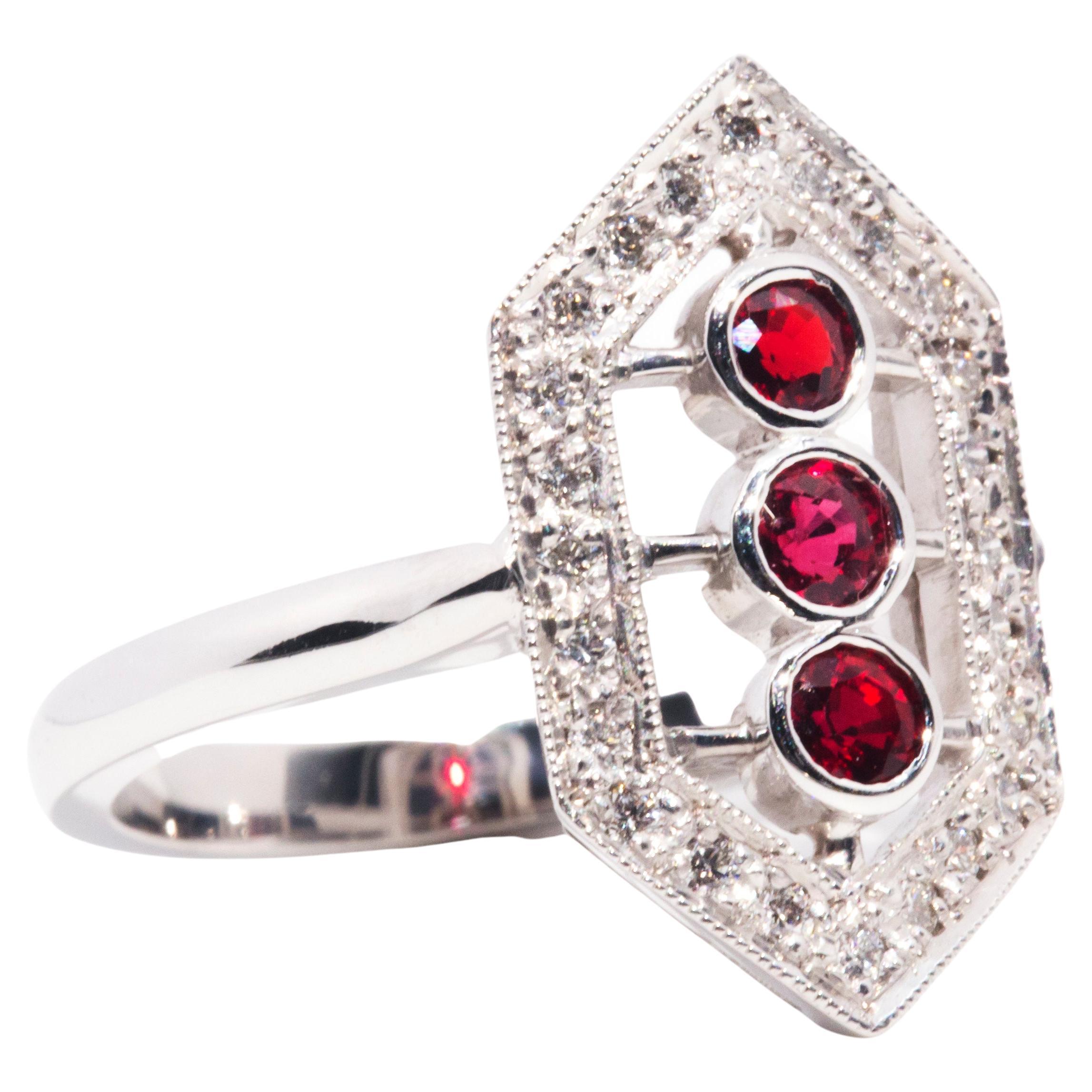 This gorgeous Art Deco inspired ring is forged in 18 carat white gold and features a breathtaking elongated hexagonal cluster embellished with an opulent border of sparkling round brilliant cut diamonds and three alluring bright red natural red
