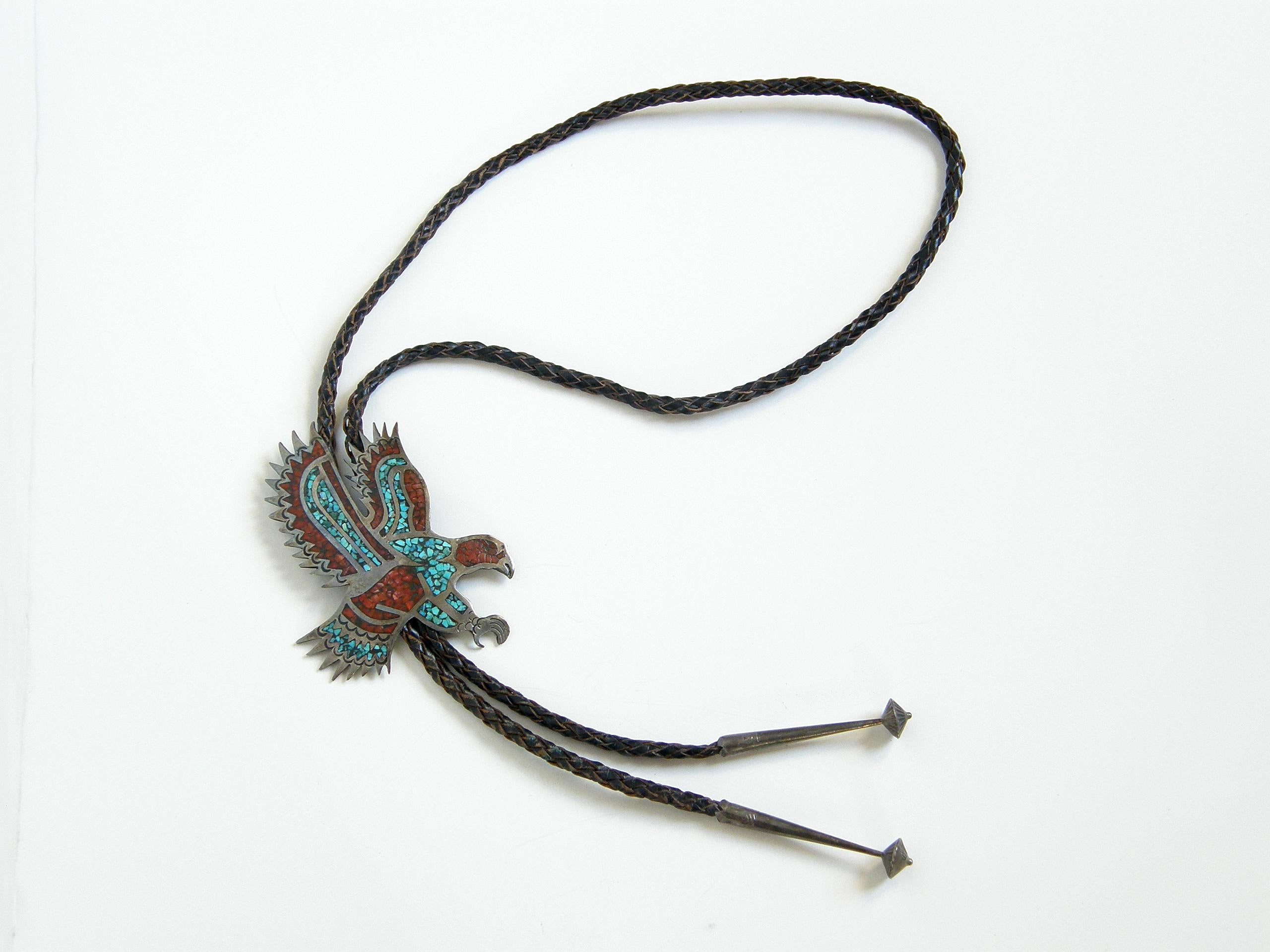 This circa 1970s, handmade bolo tie features a dramatic,  sterling eagle in flight. The silver has turquoise and coral chip inlay that enhances the design. The braided leather cord ends with decorative, sterling tips, and there is a locking slide on