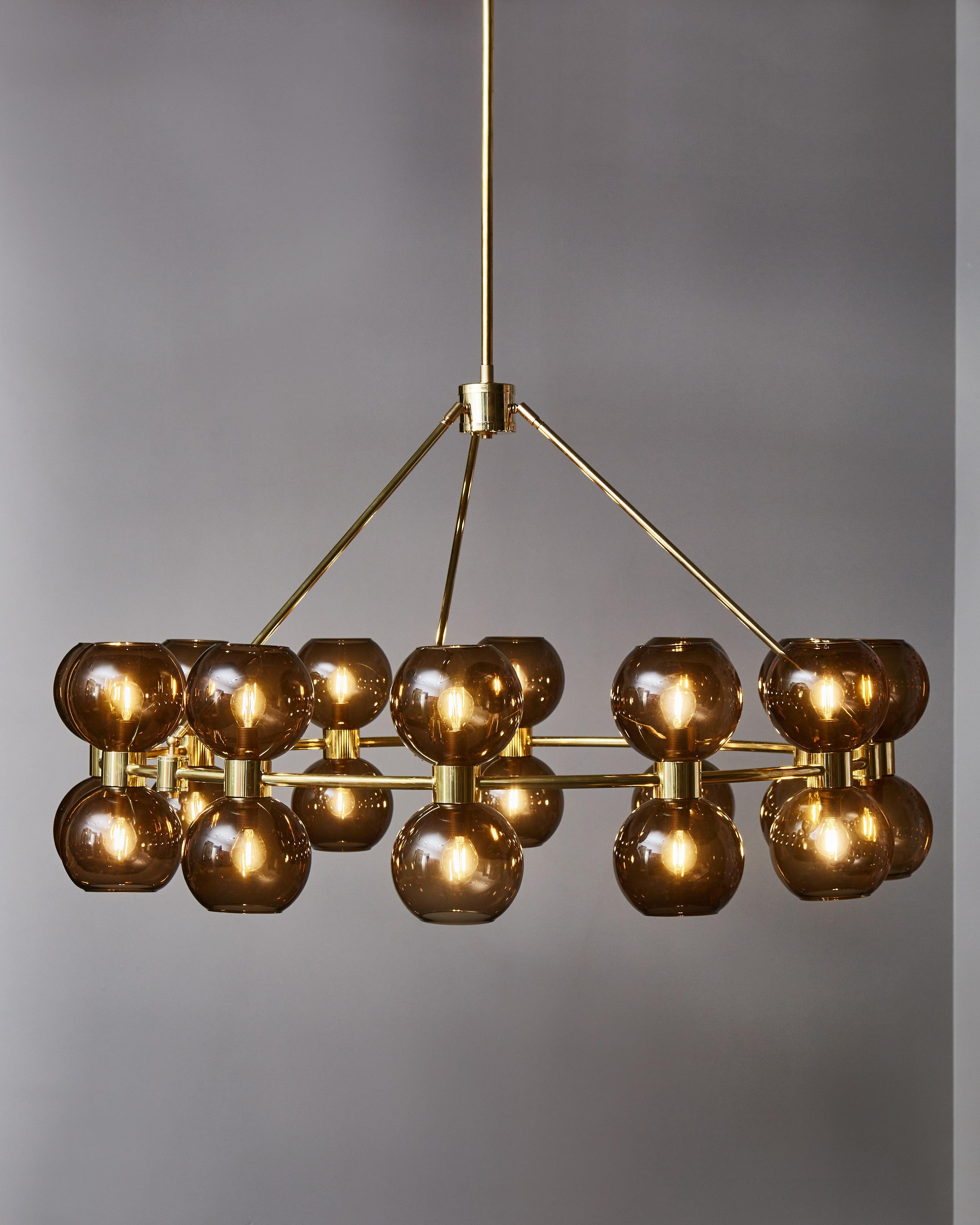 Lampadario rotondo in ottone con ventiquattro sorgenti luminose, ciascuna ricoperta da globi di vetro colorato marrone. Il cerchio principale è costituito da tre aste che si uniscono al gambo centrale.