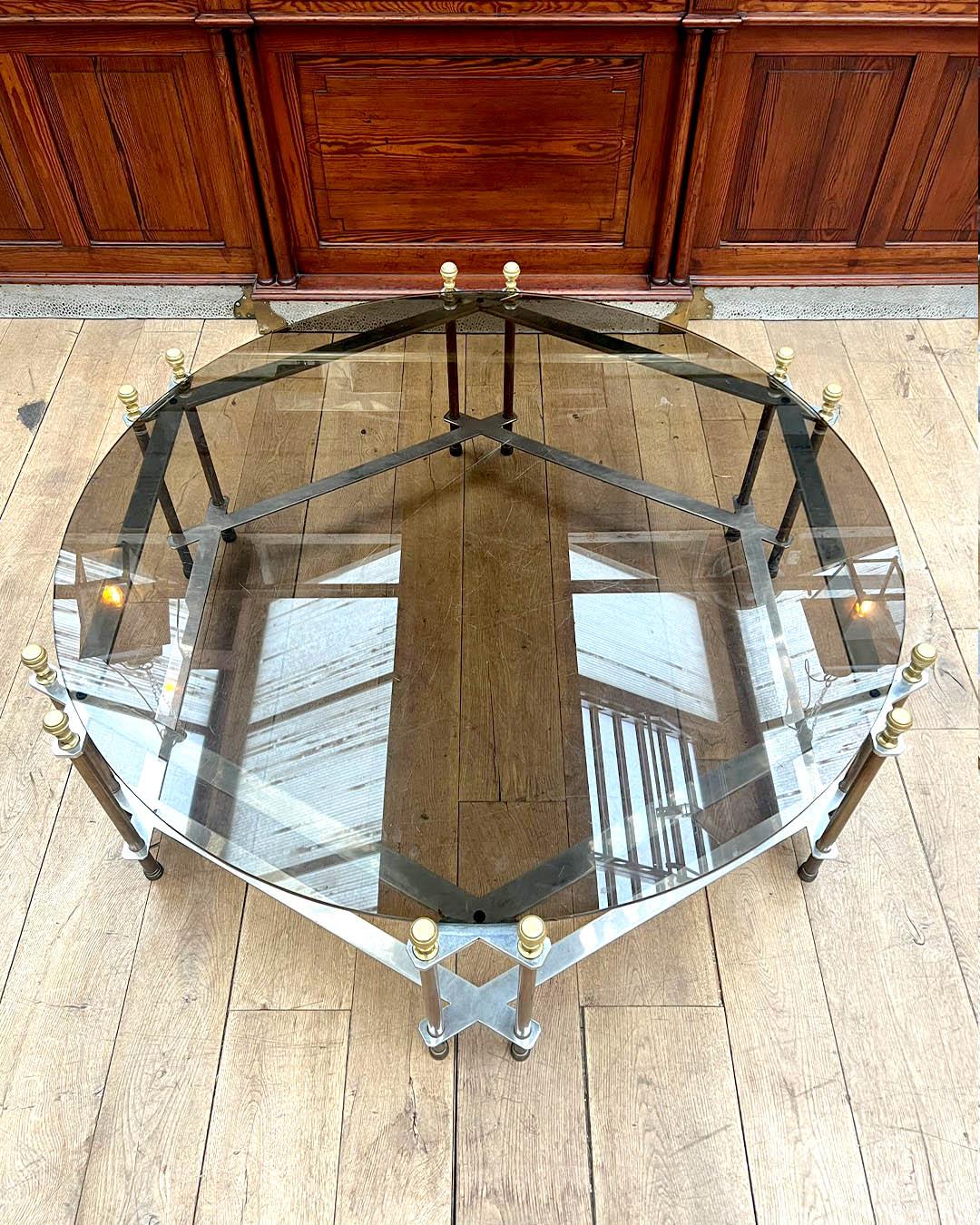 Elegant circular coffee table featuring a smoked glass top supported by a gilt bronze and steel frame with decorative finials. The table combines modernist lines with classical detailing, reflecting the glamorous design aesthetic of the 1970s French