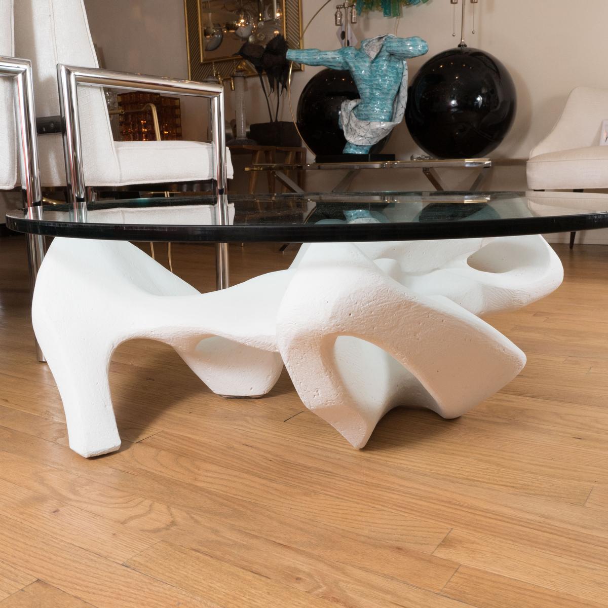 Circular coffee table with white composite sculptural base and glass top. 
