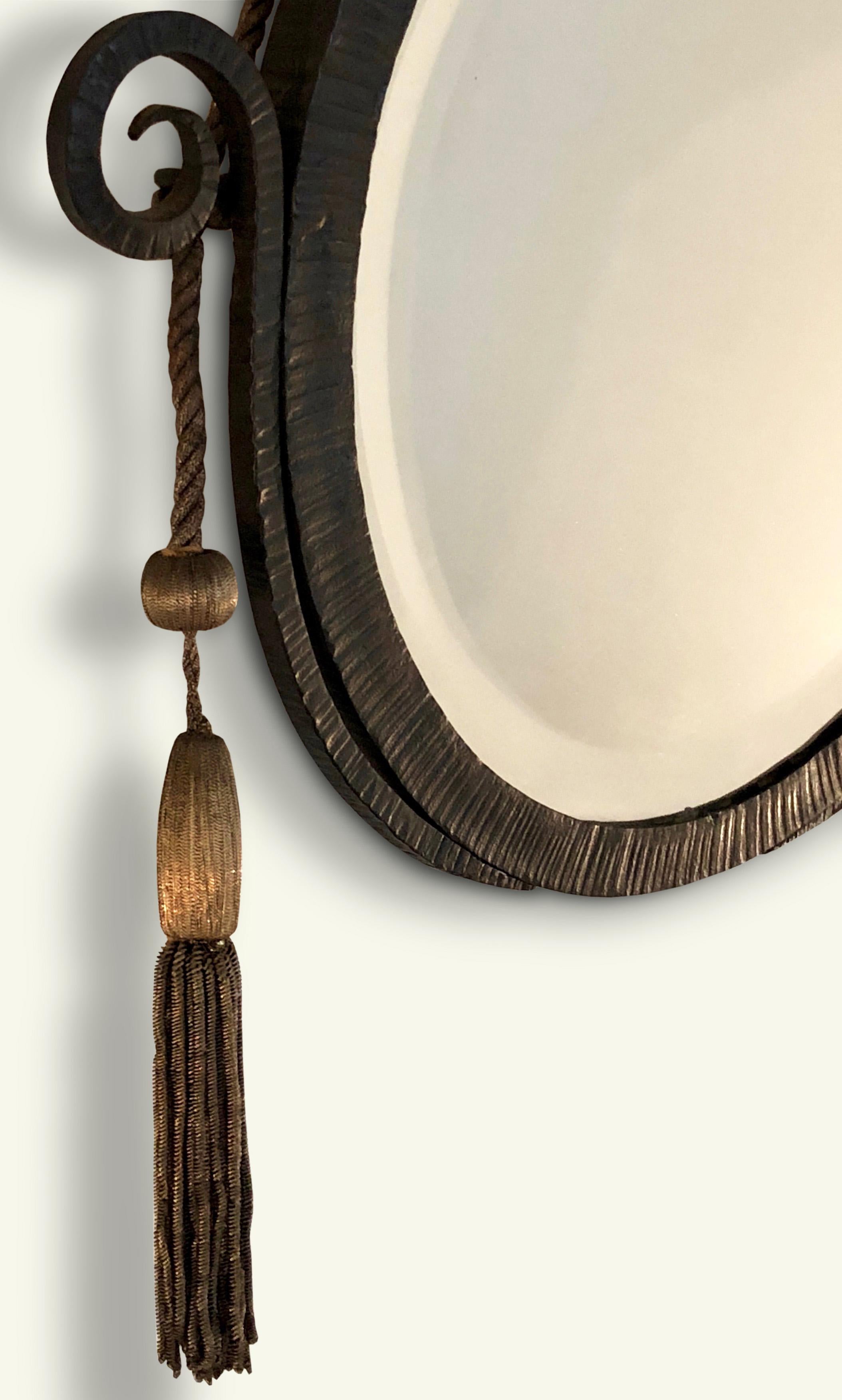 An elegant circular Art Deco Mirror in the manner of Edgar Brandt. The ironwork frame features a textured ridged edge typical of his work with a decorative scrolled motif at either side. The mirror hangs from a decorative woven metallic tasseled