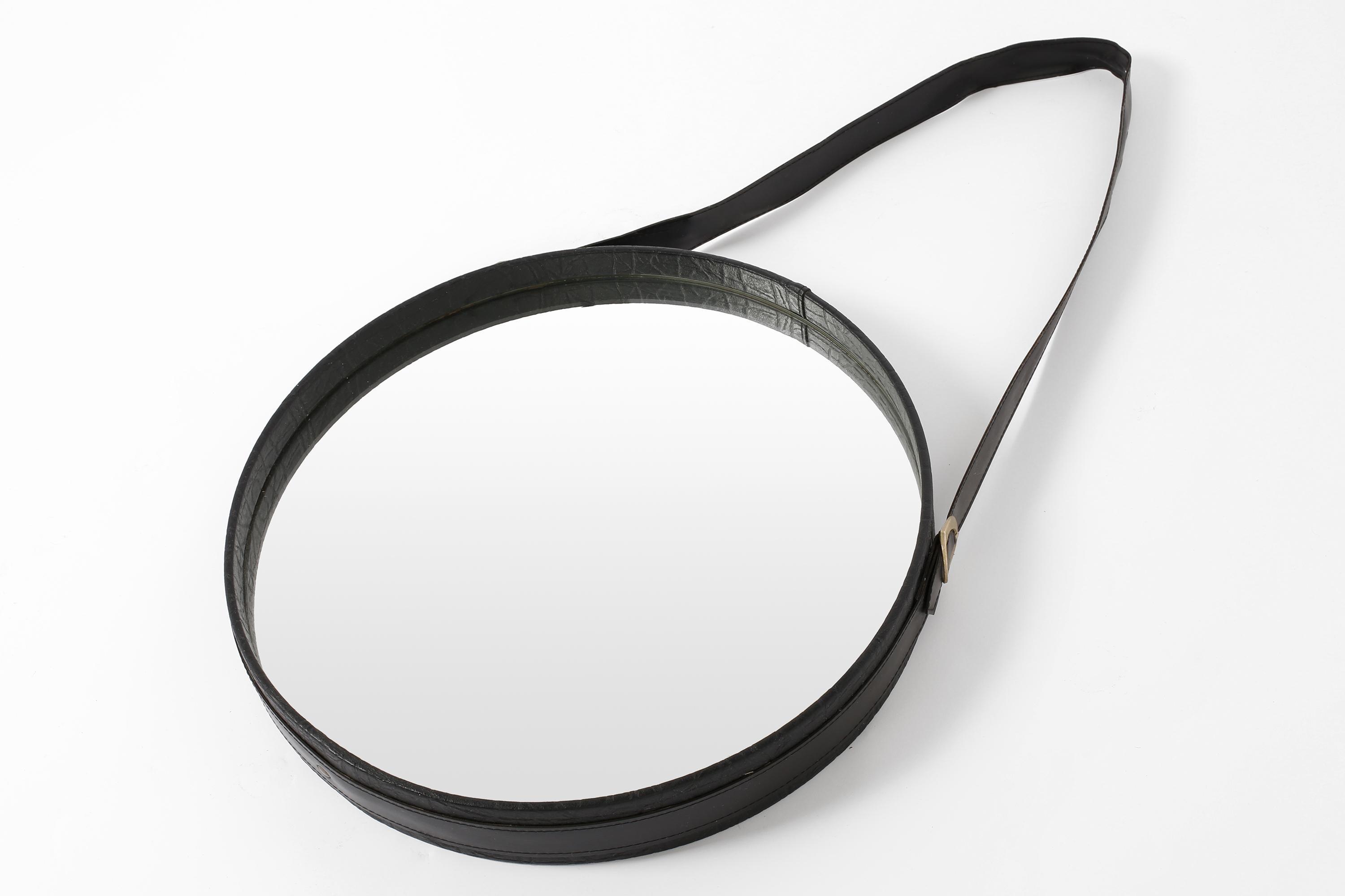 A circular hanging wall mirror, clad in black faux leather with brass hardware. French, c. 1970s.