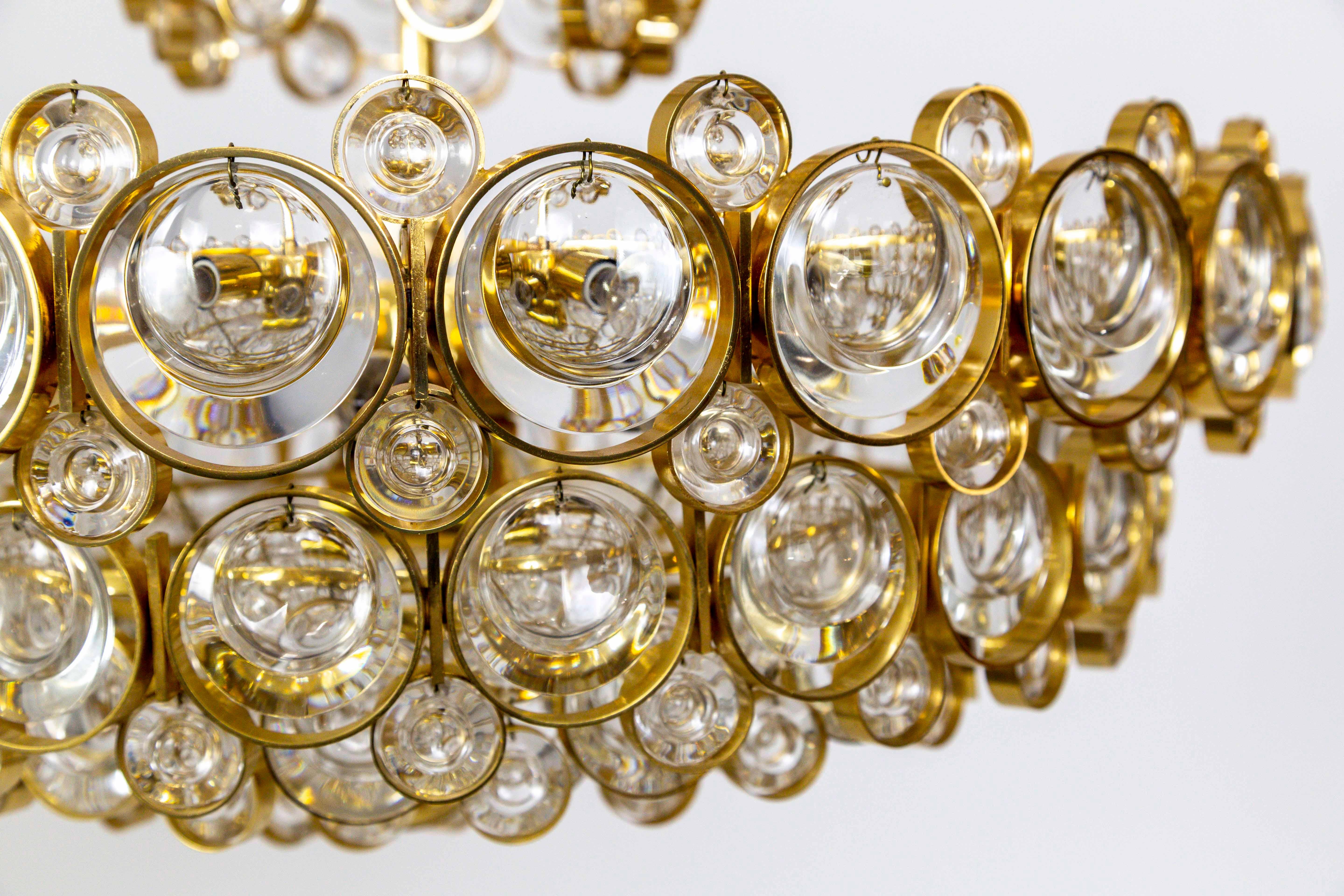 A Mid-Century Modern Palwa chandelier with a complex, multi tier design with 11 lights; a gilded brass, circular structure comprised of small and large rings with dangling optical glass lens crystals inside. 5 candelabra and 6 medium base, porcelain