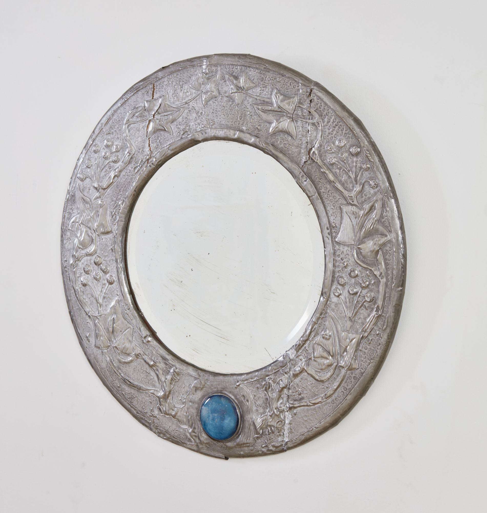 An Arts and Crafts period circular mirror with wide pewter frame having embossed decoration of a winding grapevine with hand-hammered punchwork detailing, with central beveled mirror plate, and a large blue cabuchon at the base. Can easily be