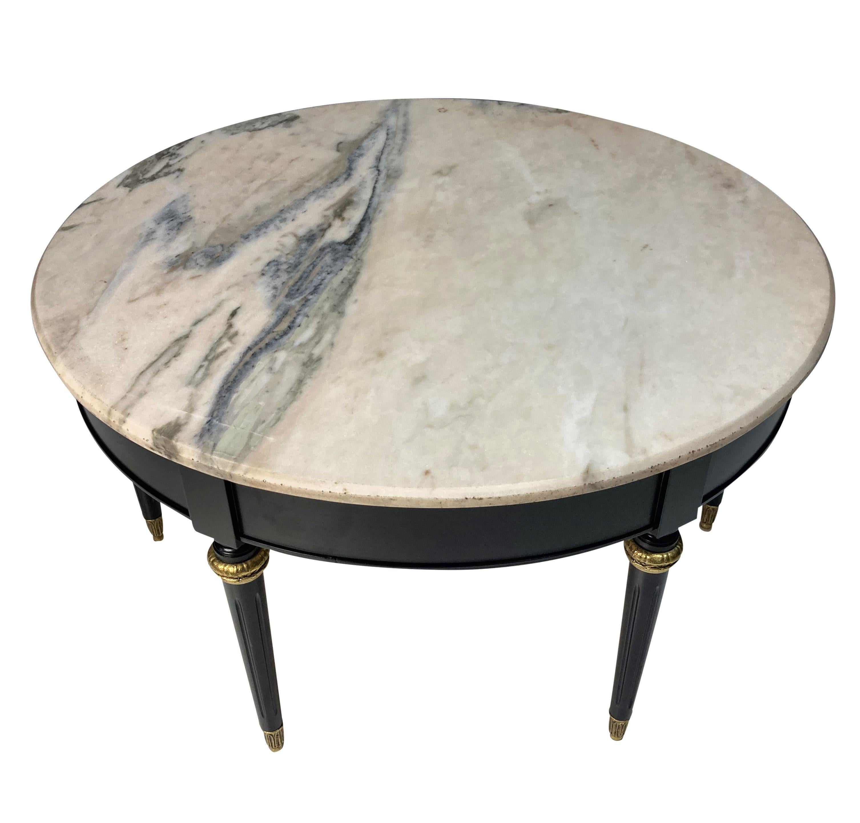A French circular occasional table by Maison Jansen, ebonised, with six tapering fluted legs with gilt brass sabot and collars. The variegated marble top is creamy ink with a grey vein.
