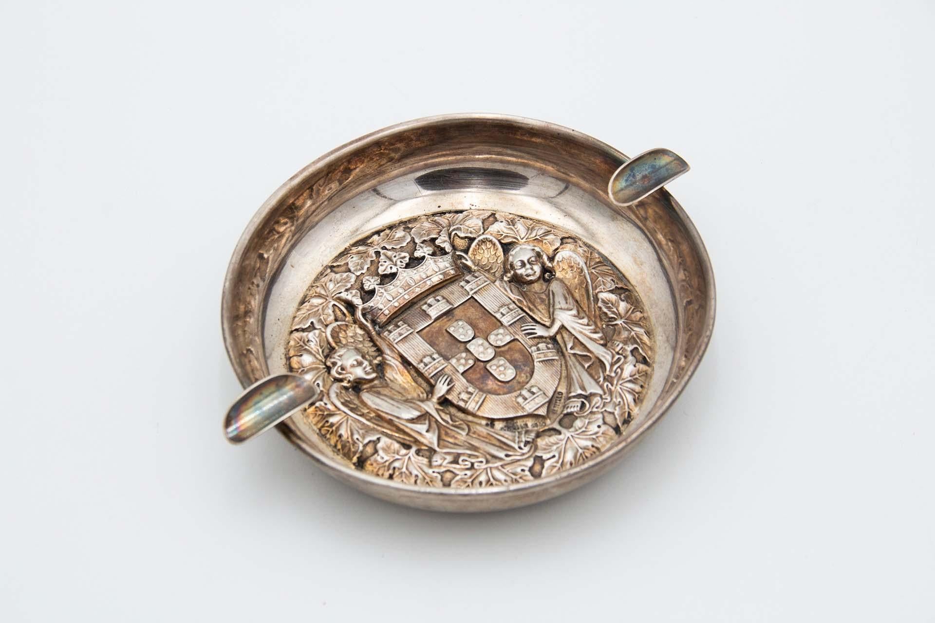 Description : Cendrier circulaire en argent avec deux porte-cigarettes et les armoiries du Portugal avec deux anges. Métal : Argent. Origine : Portugais. Marque : Eagle Head - I+I Titre. Poids : 131,74 grammes. Dimensions : 17 cm x 3,3 cm :