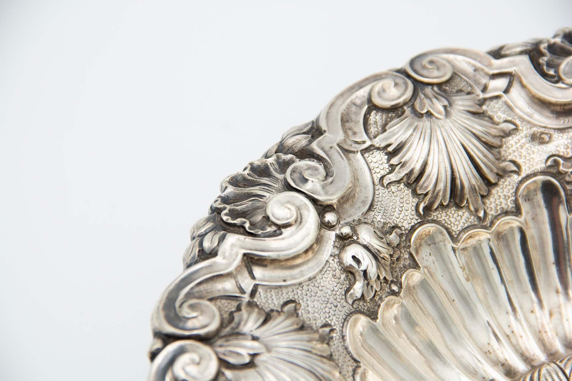 Description: Circular silver fruit bowl centerpiece profusely embossed with shells and flutes. Metal: Silver. Origin: Portuguese. Brand: javali II Title. Weight: 274.80 grams. Dimensions: 23.6cmx8.3cm. Content: 833/1000. Assay Mark: Yes, between