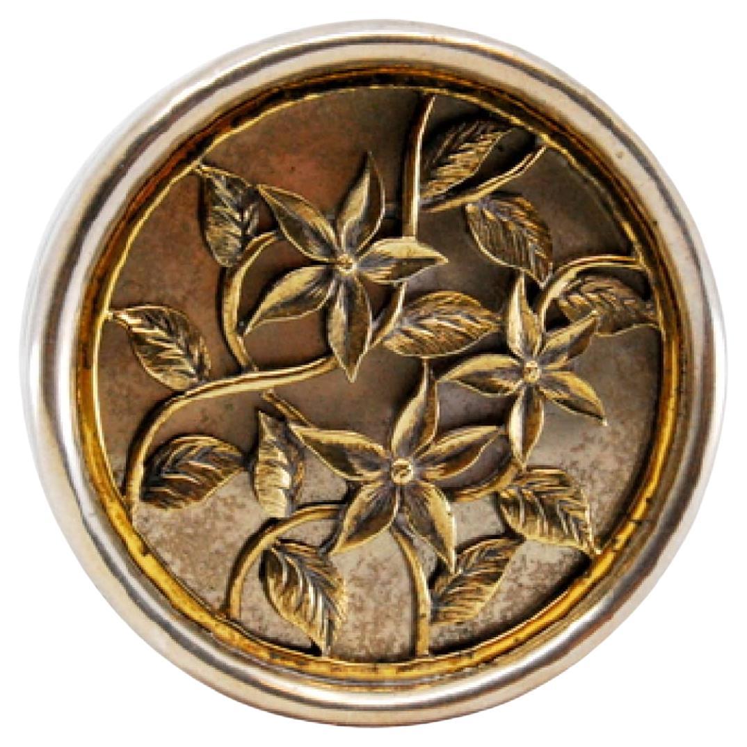 Circular silver pill box with gold floral motifs embossed on the removable lid