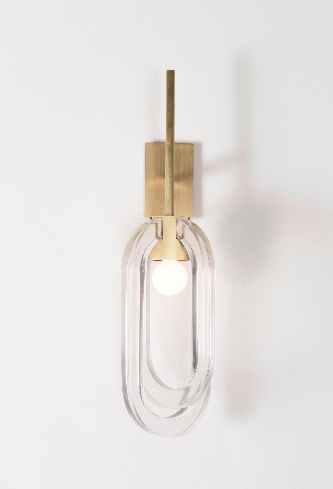 Two cast-resin rings, placed perpendicularly around a brushed-brass light source, each at varied lengths with one looping under the other to create a composed, jewel-like fixture. The fittings and mount are made of machined brass a fogged -dome