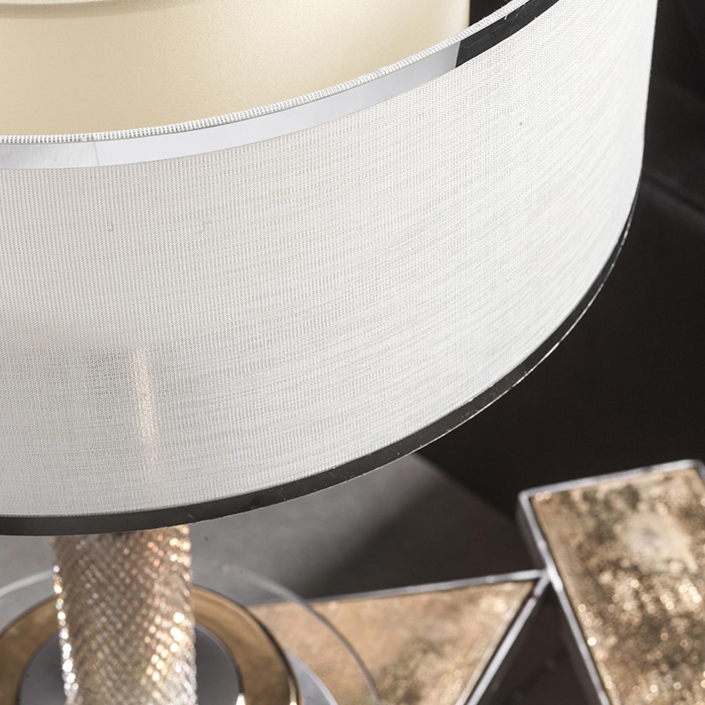 This table lamp features a nickel-plated brass stem covered with crystal details, while its base has rounded accents in gold-plated majolica pottery. A horizontal shade adds visual and dimensional complexity, with a silver, woven-fabric exterior and