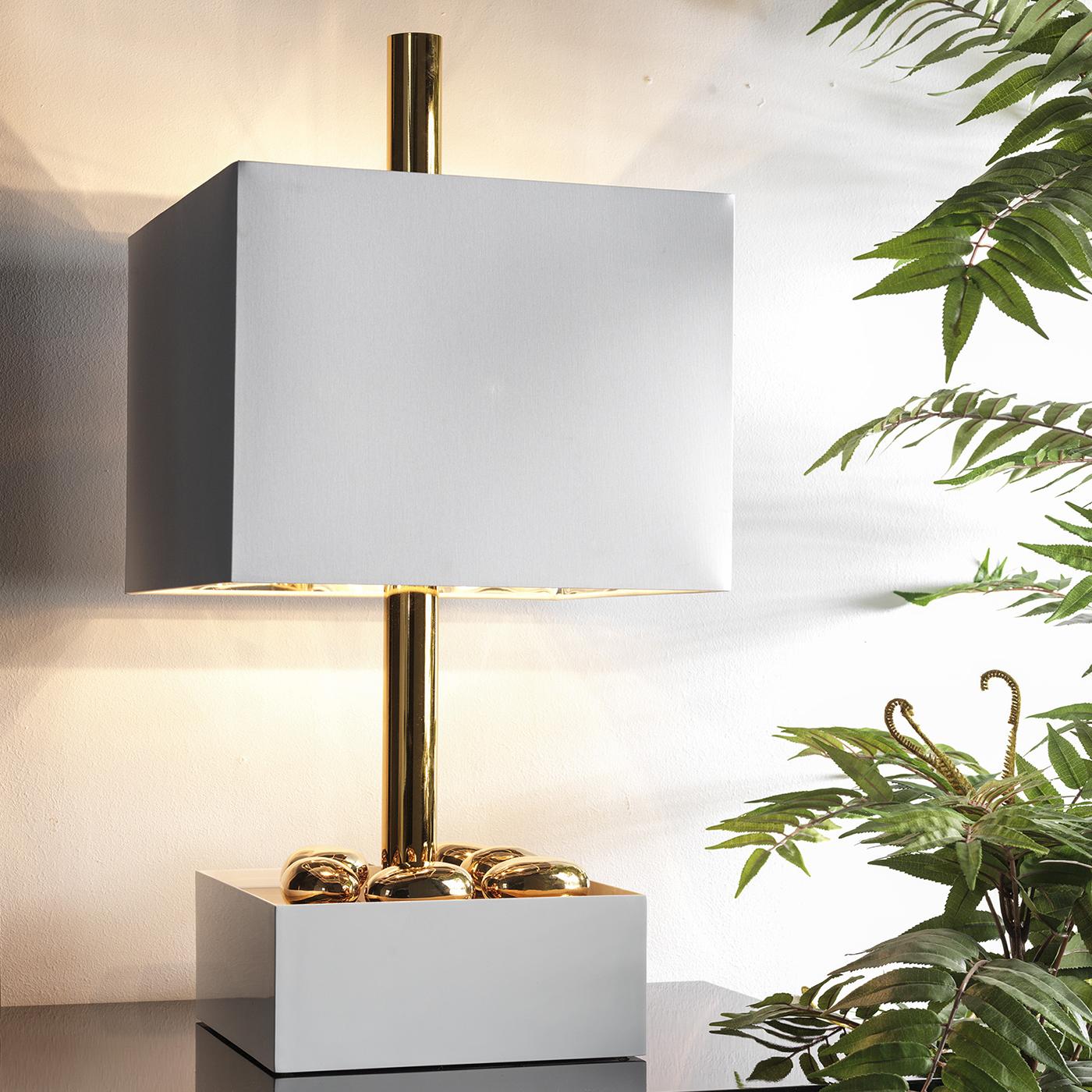 This table lamp's flat planes create the illusion of lacking dimensionality. The opaque whiteness of its shade and base contrast with its lustrous gold-plated stem, creating a surprising burst of shine and color. The style is completed by a chintz