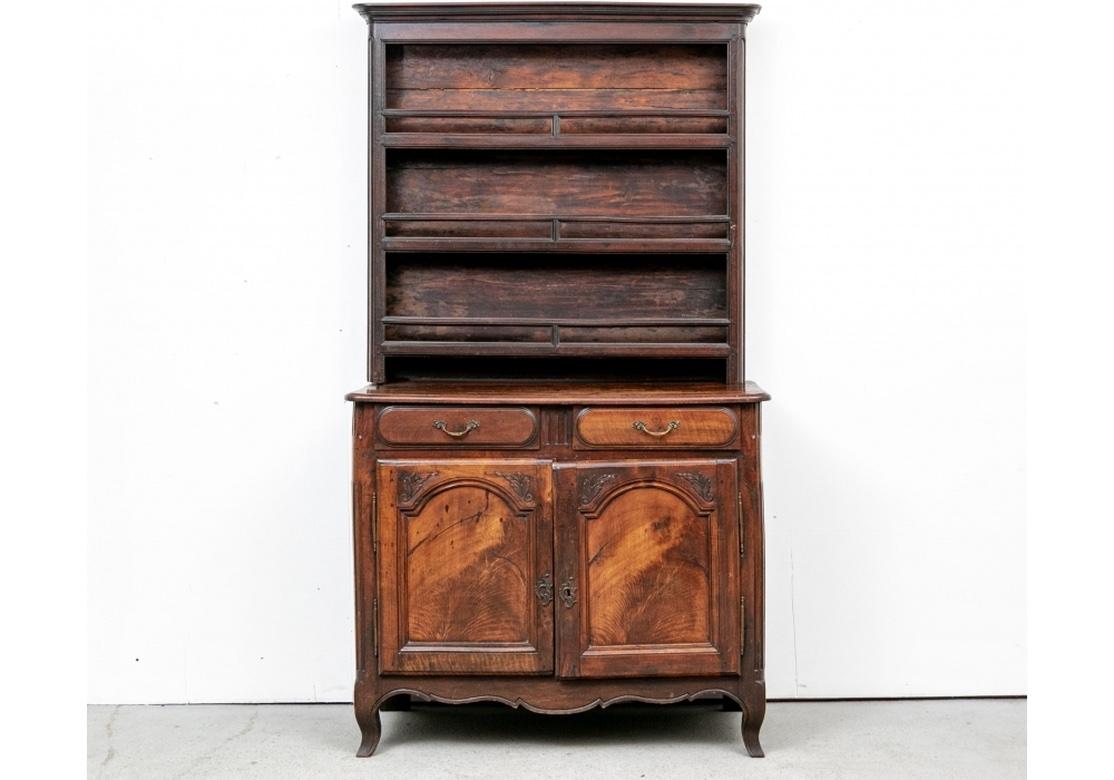 A two-part hutch. The top with a carved cornice over three shelves for plates with carved moldings. The lower cabinet with deep top over two short frieze drawers with oval moldings and fine brass foliate pulls. The figured wood double doors with