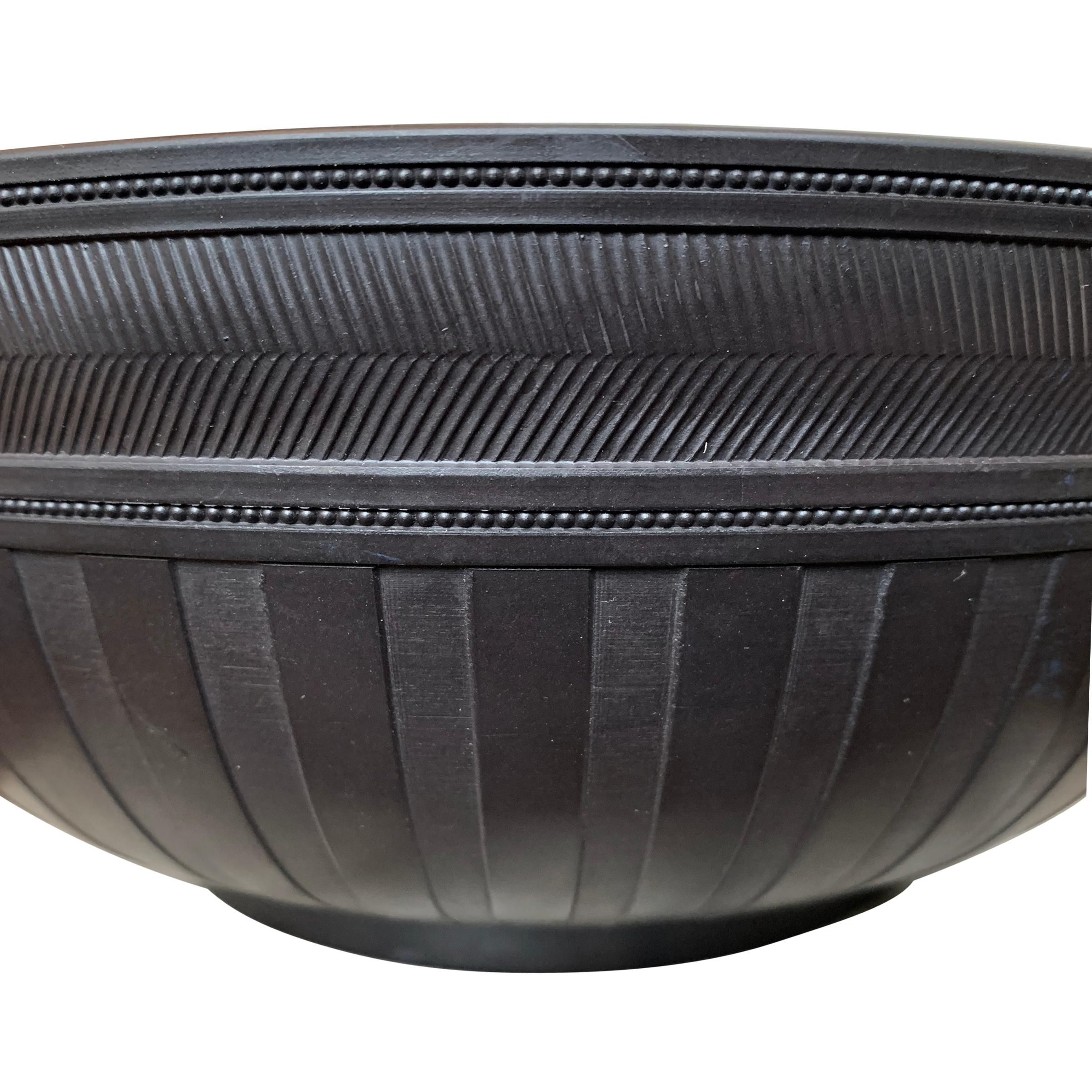 A 1967 re-edition of a classic 19th century Wedgwood basalt ceramic bowl with a horizontal herringbone and beaded pattern band at the lip, vertical machine-cut stripes around the belly, and raised on a short foot. This pattern dates to the early